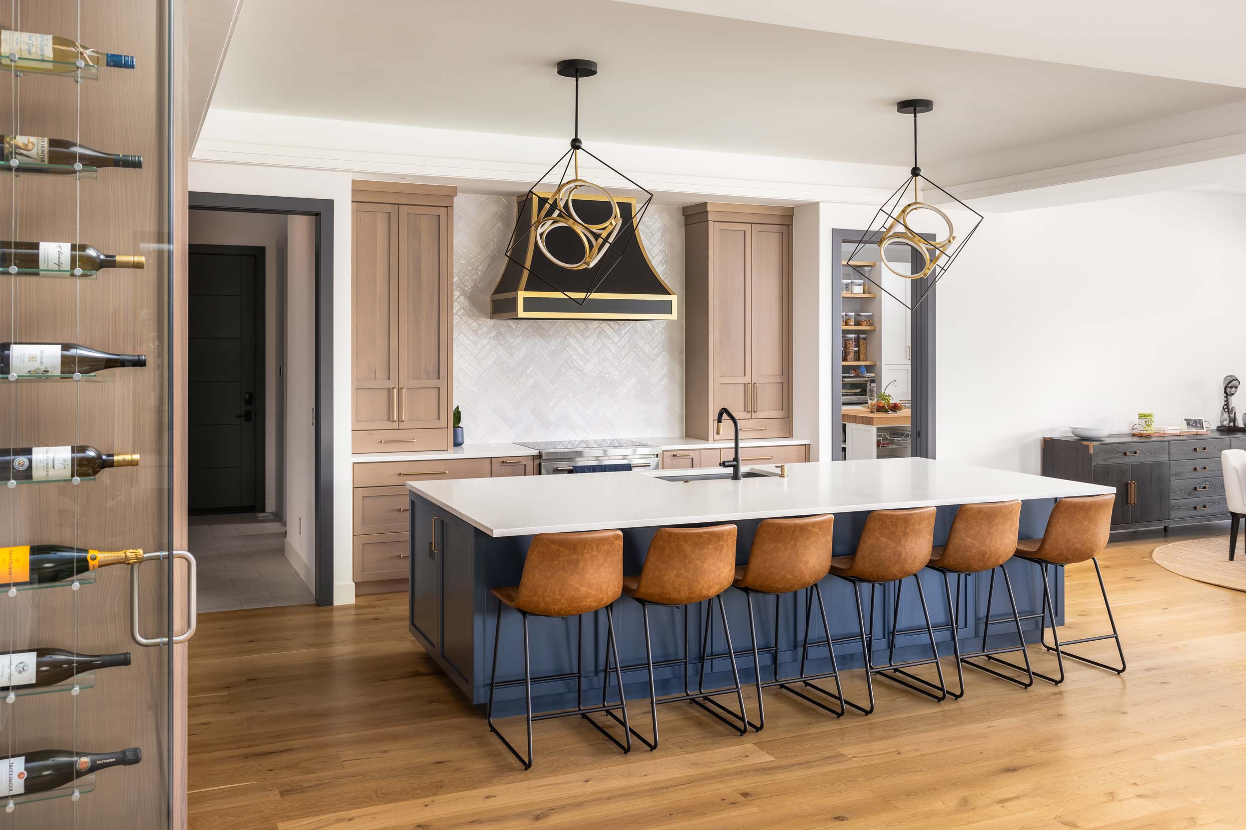 Modern kitchen with island, four brown bar stools, pendant lights, and built-in shelves, with wooden flooring and white walls.
