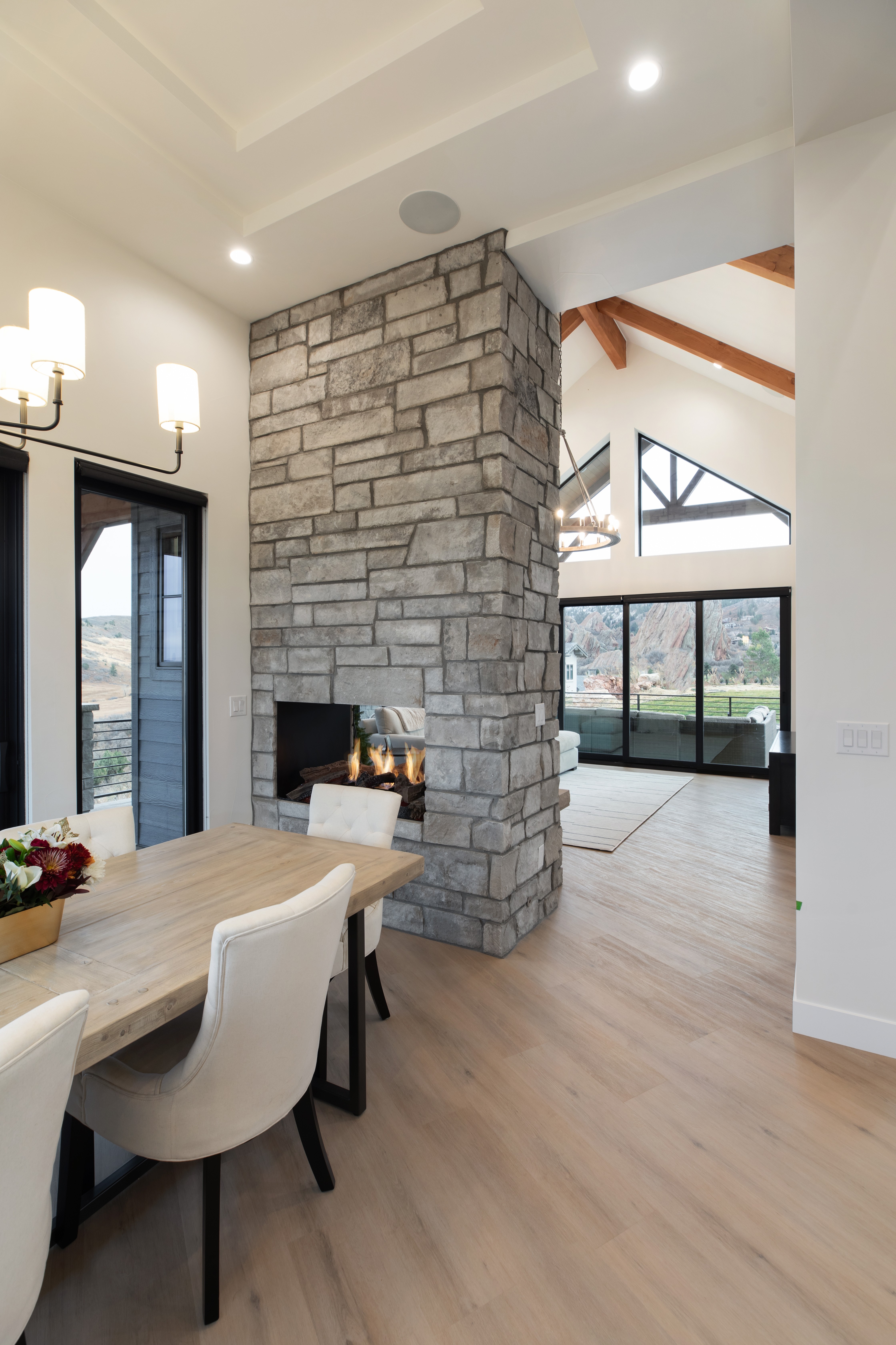 Interior of a modern living space with a stone fireplace, dining area, and large windows showing outdoor scenery.