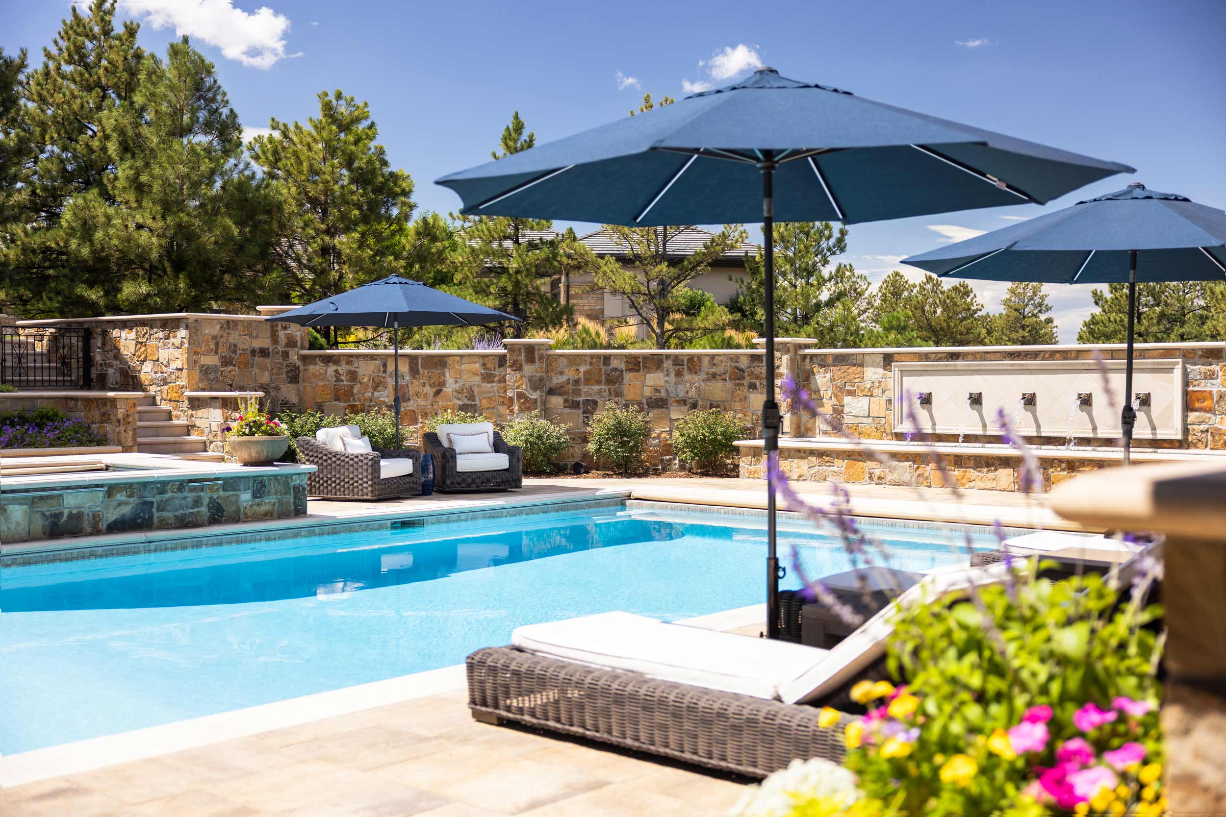 Swimming pool with lounge chairs and umbrellas outdoors, surrounded by trees and a stone wall.