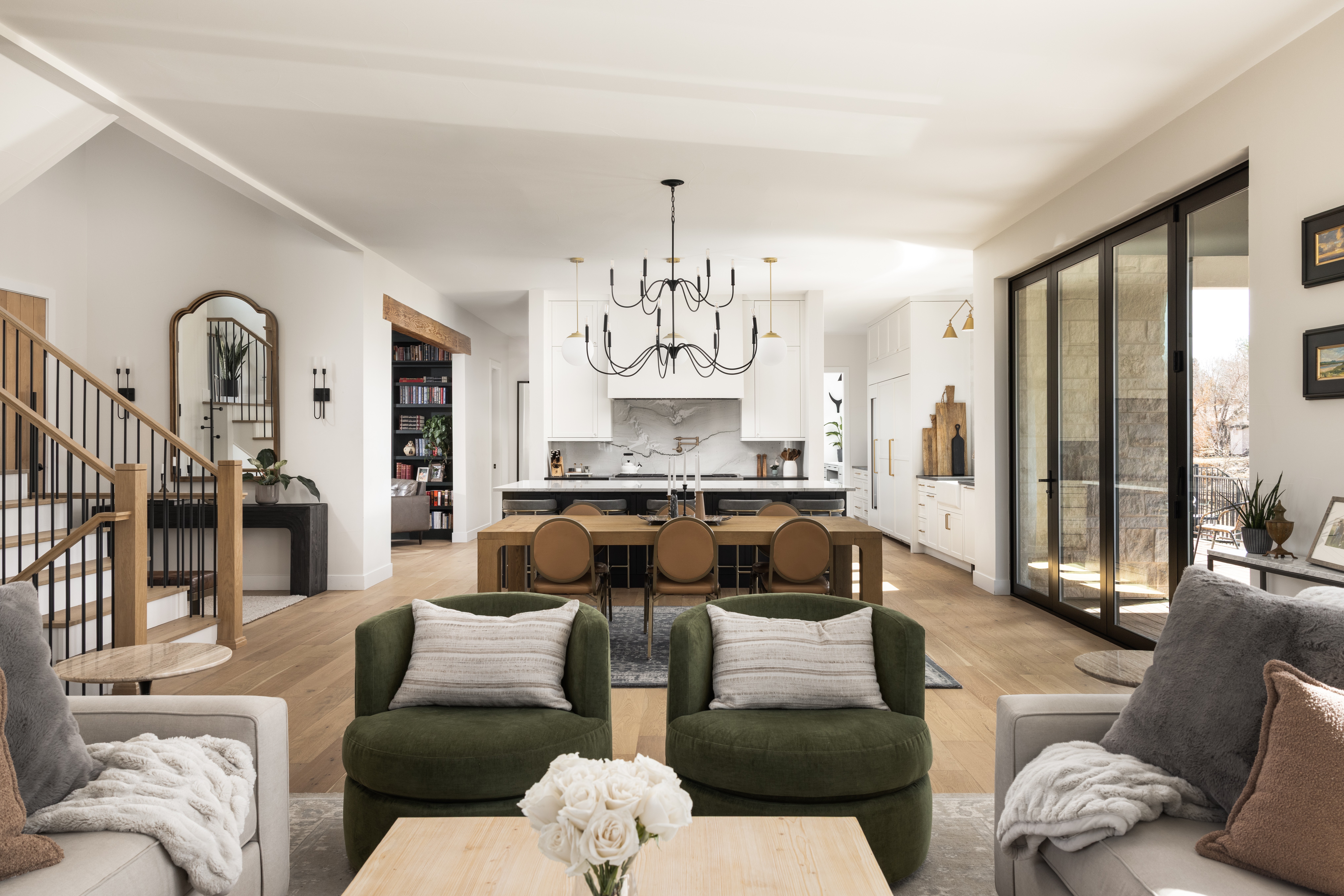 Living room with two green armchairs, a white sofa, and a dining area with a chandelier, open kitchen, and large sliding glass doors.