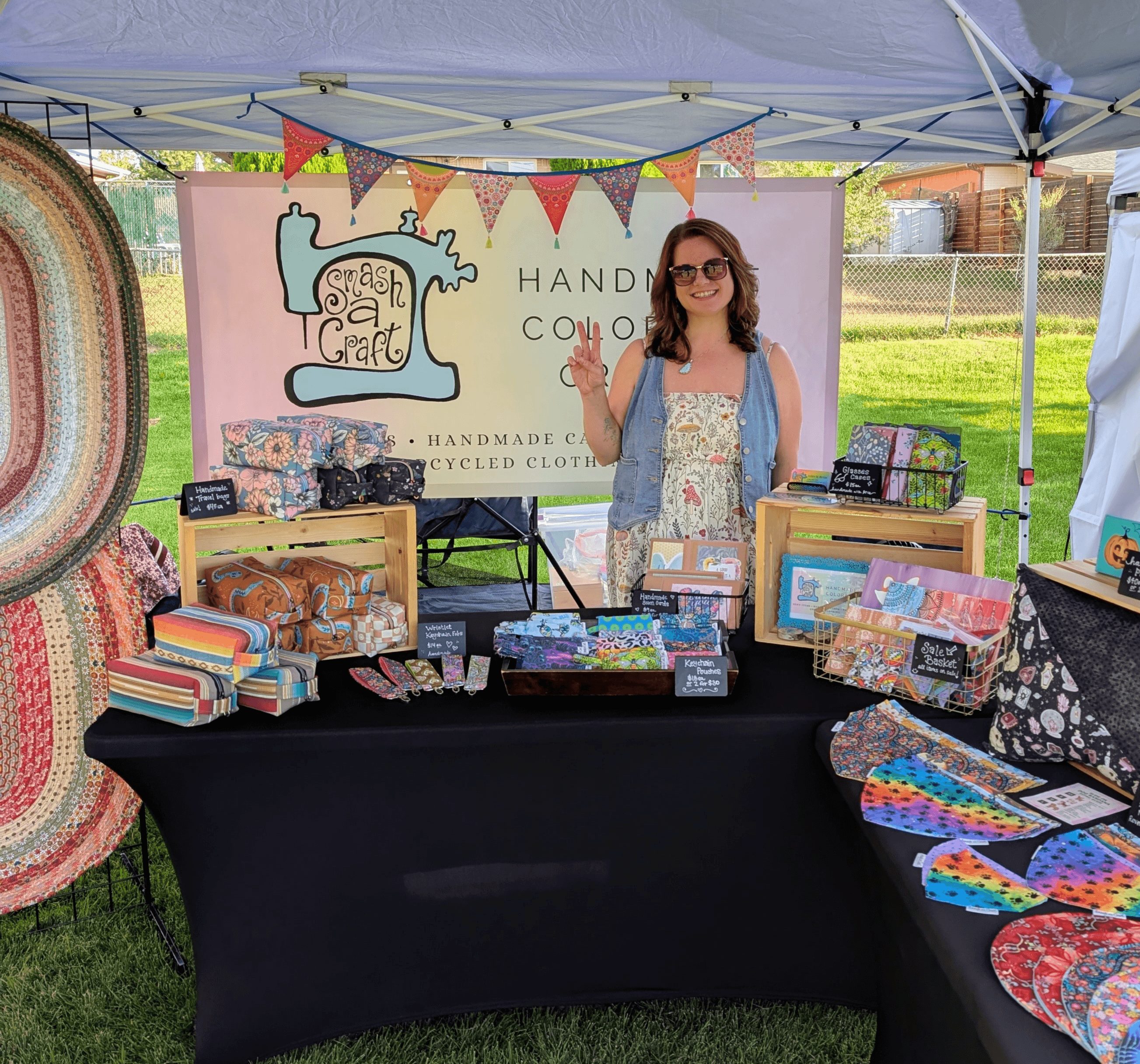 Woman standing behind a craft booth with handmade items, outdoors under a canopy, smiling and waving.