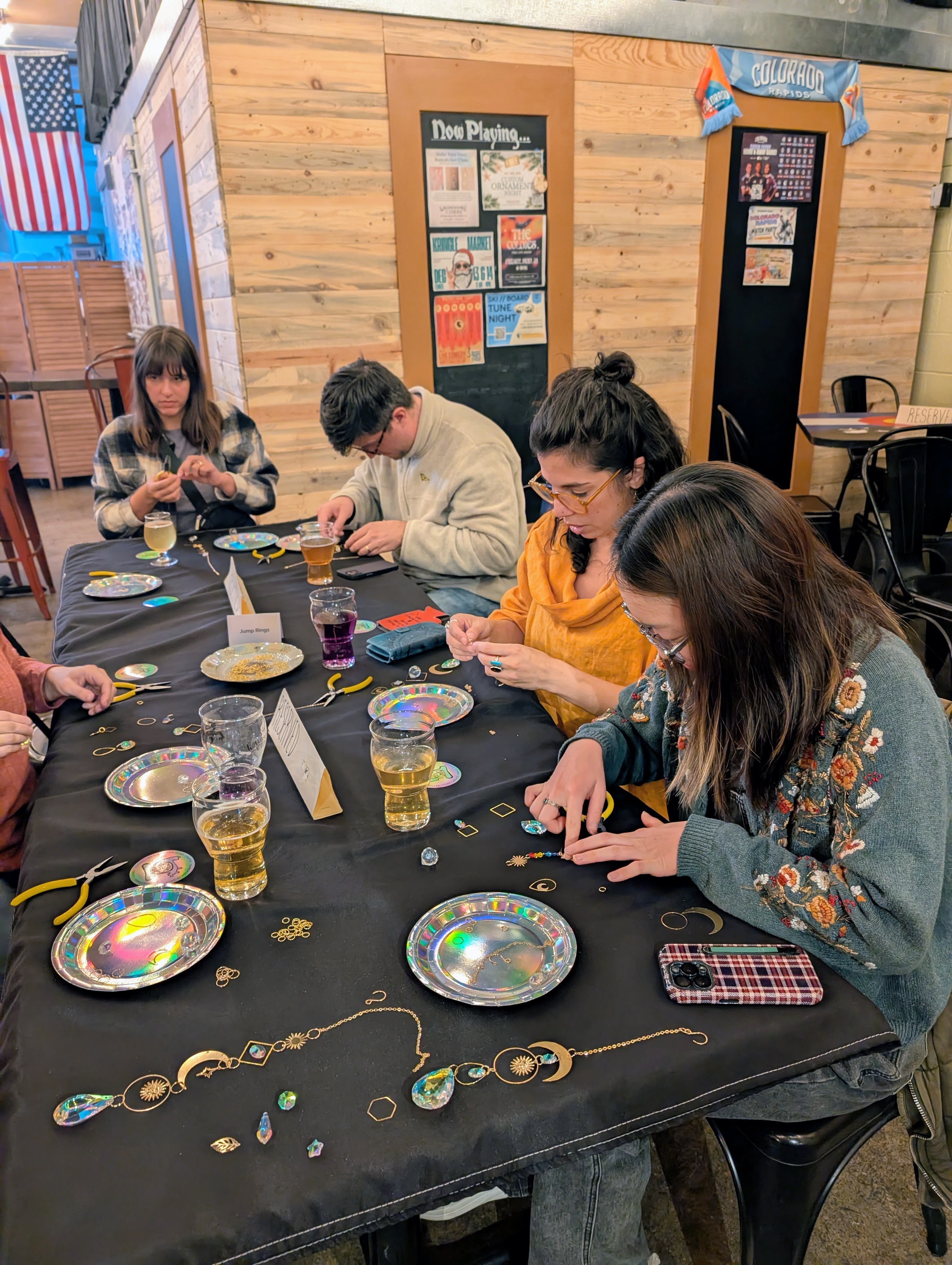 Group of four people sitting at a table with jewelry and drinks, engaging in crafting activities indoors.