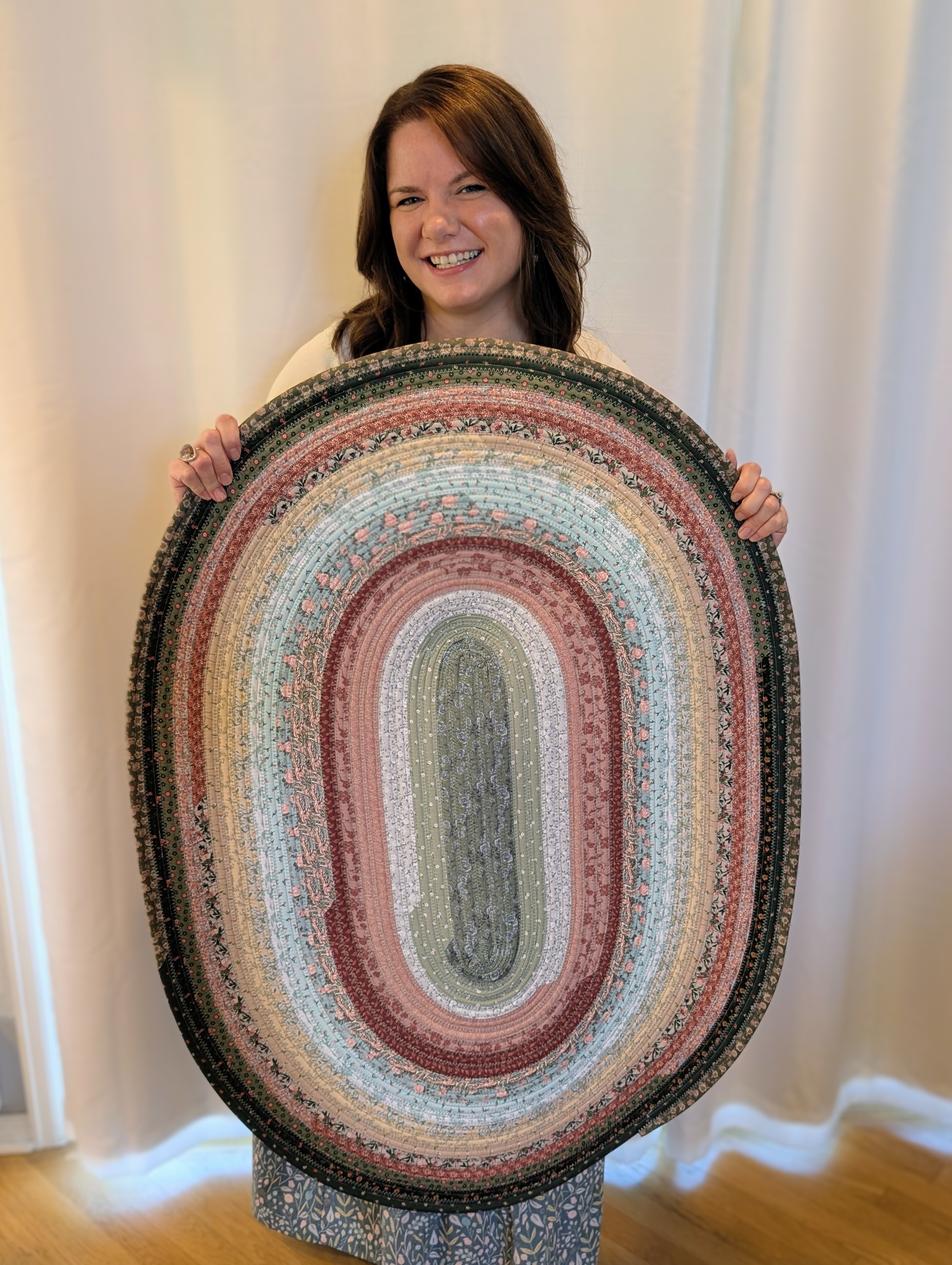 Woman holding a large oval multicolored rug, smiling, standing in front of a white curtain.
