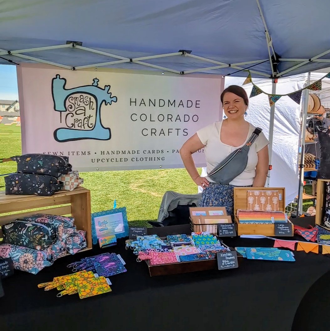 Woman standing behind a craft booth with handmade items, bags, and colorful fabrics displayed on the table, outdoors under a tent.