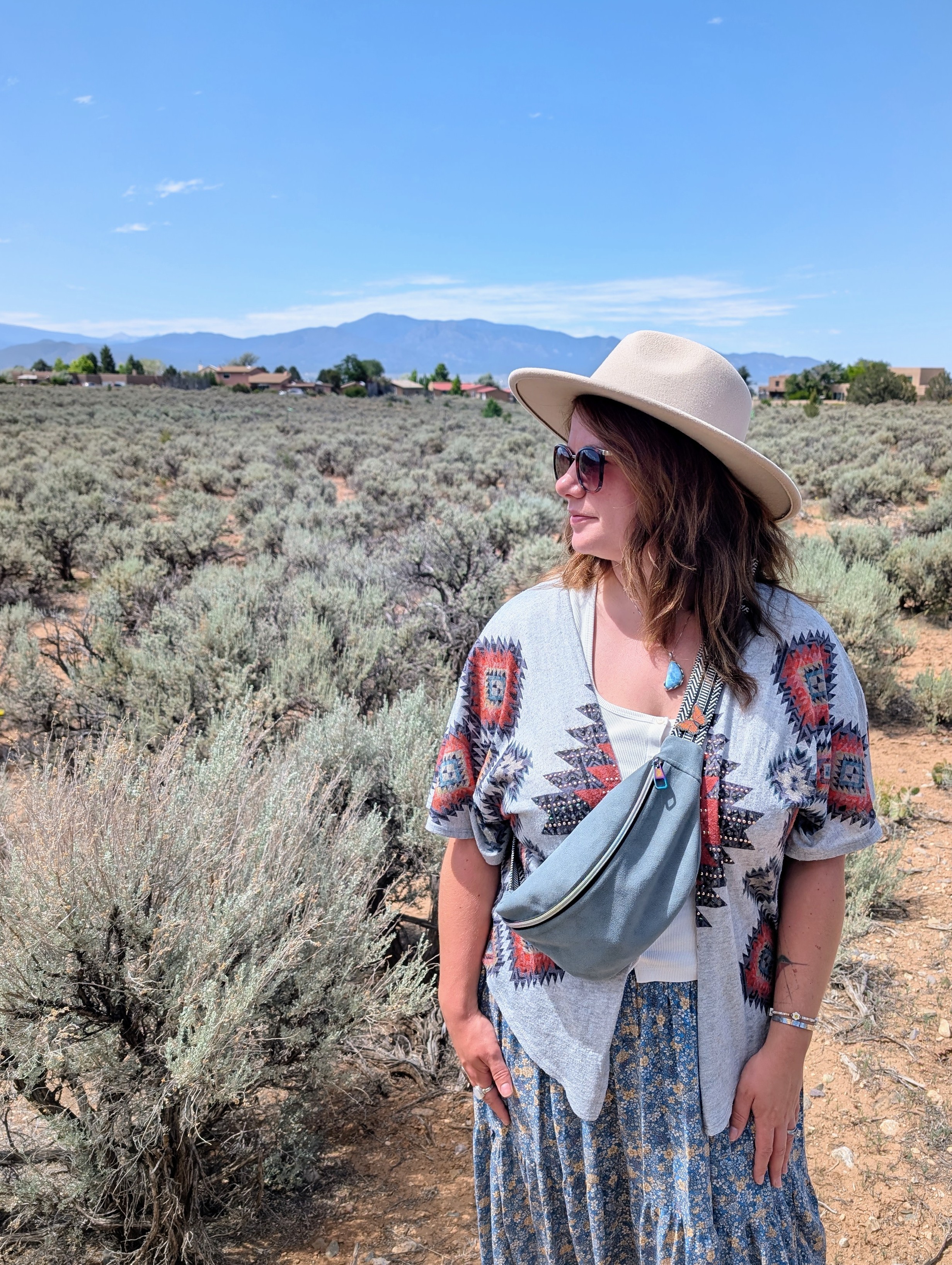 Woman wearing a hat and sunglasses standing in a desert landscape with mountains in the background.