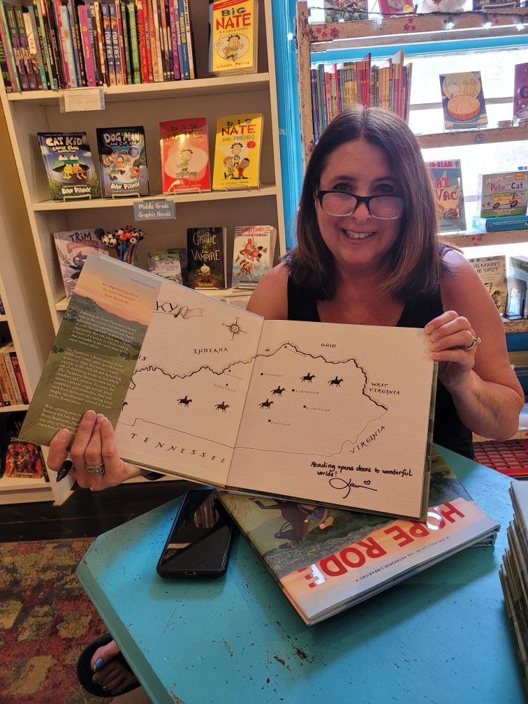 Woman with glasses smiling holding open children's book with illustrations of stars and a map, seated at a table in a bookstore.