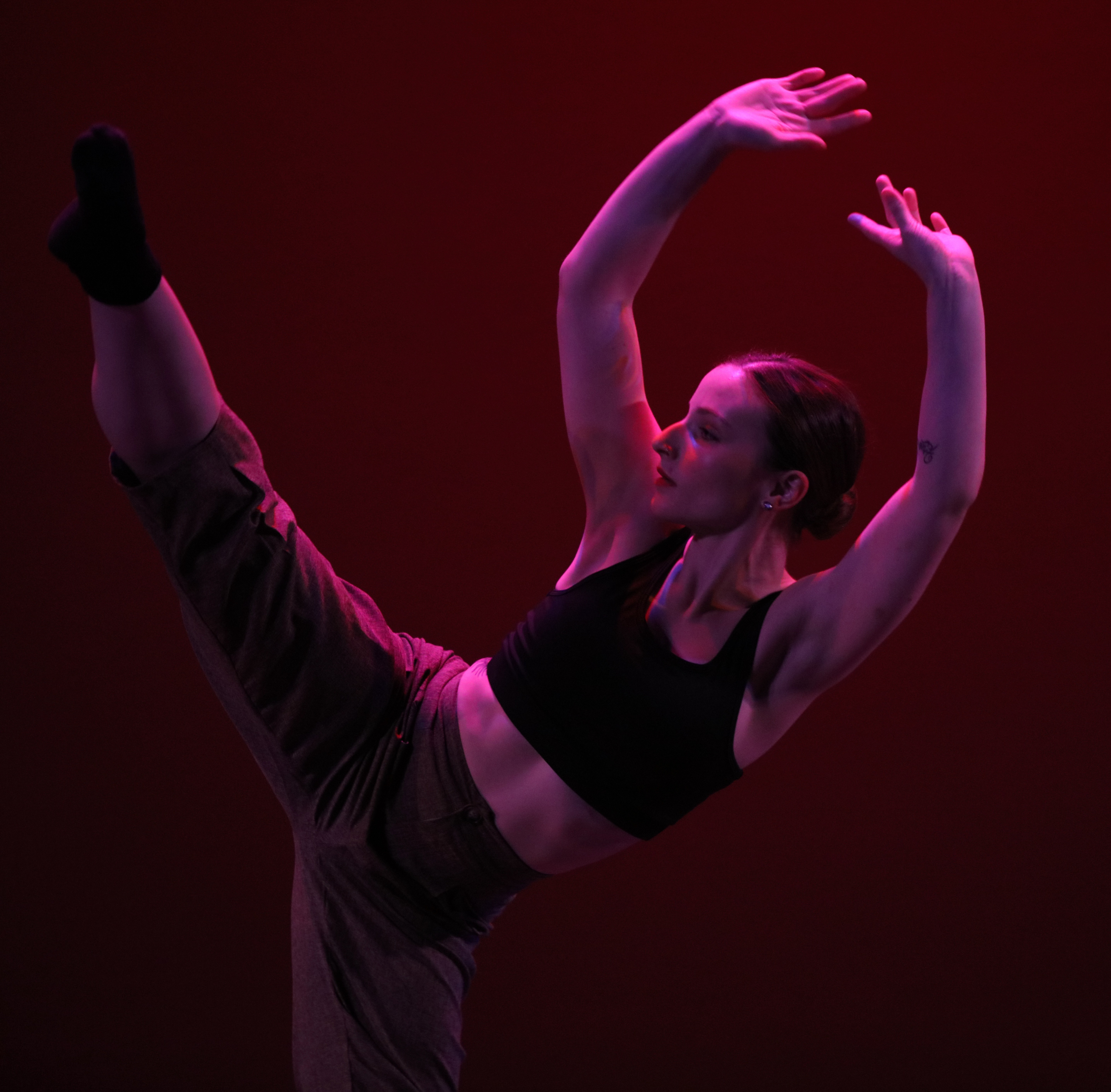 Dancer in black sports bra and pants performs a high kick with arms raised, against a dark red background.
