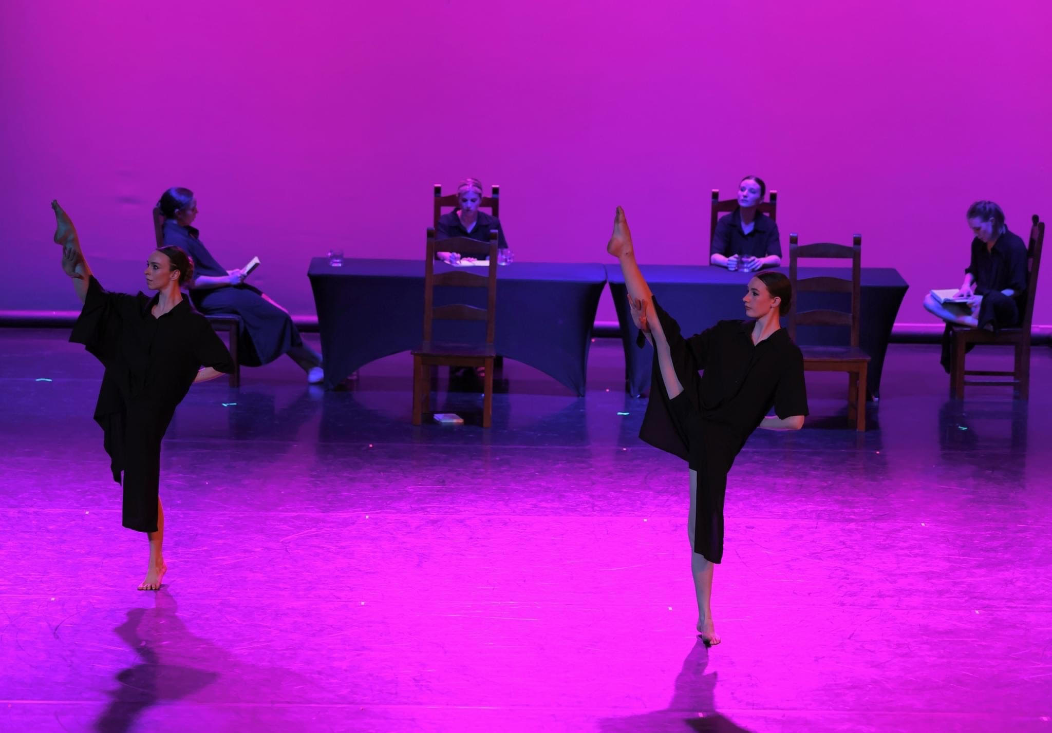 Four dancers perform on stage with purple lighting, two in front and two seated at tables in the background.