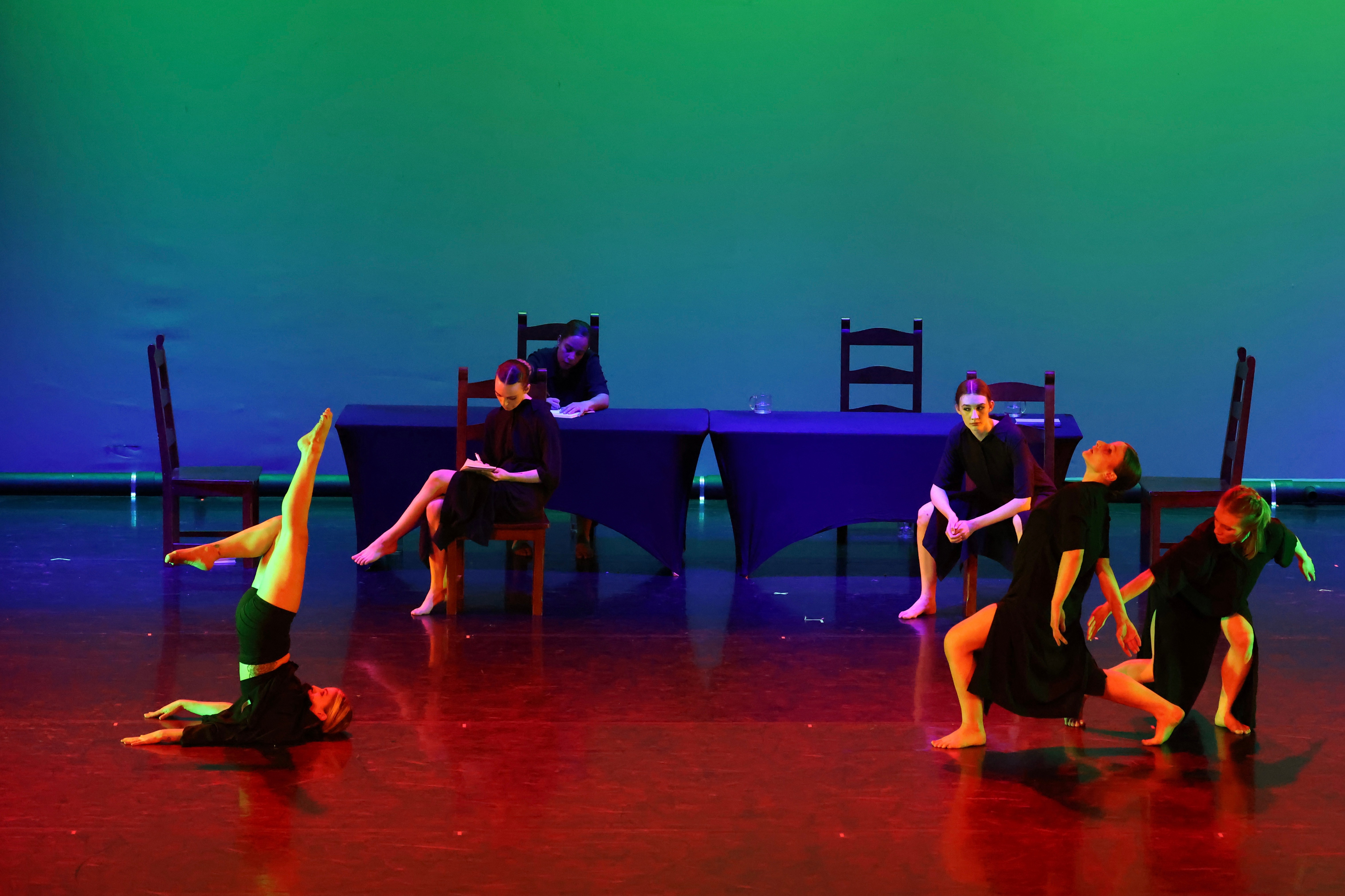 Group of five dancers performing on stage with colorful background, some in seated positions, others standing or on the floor.