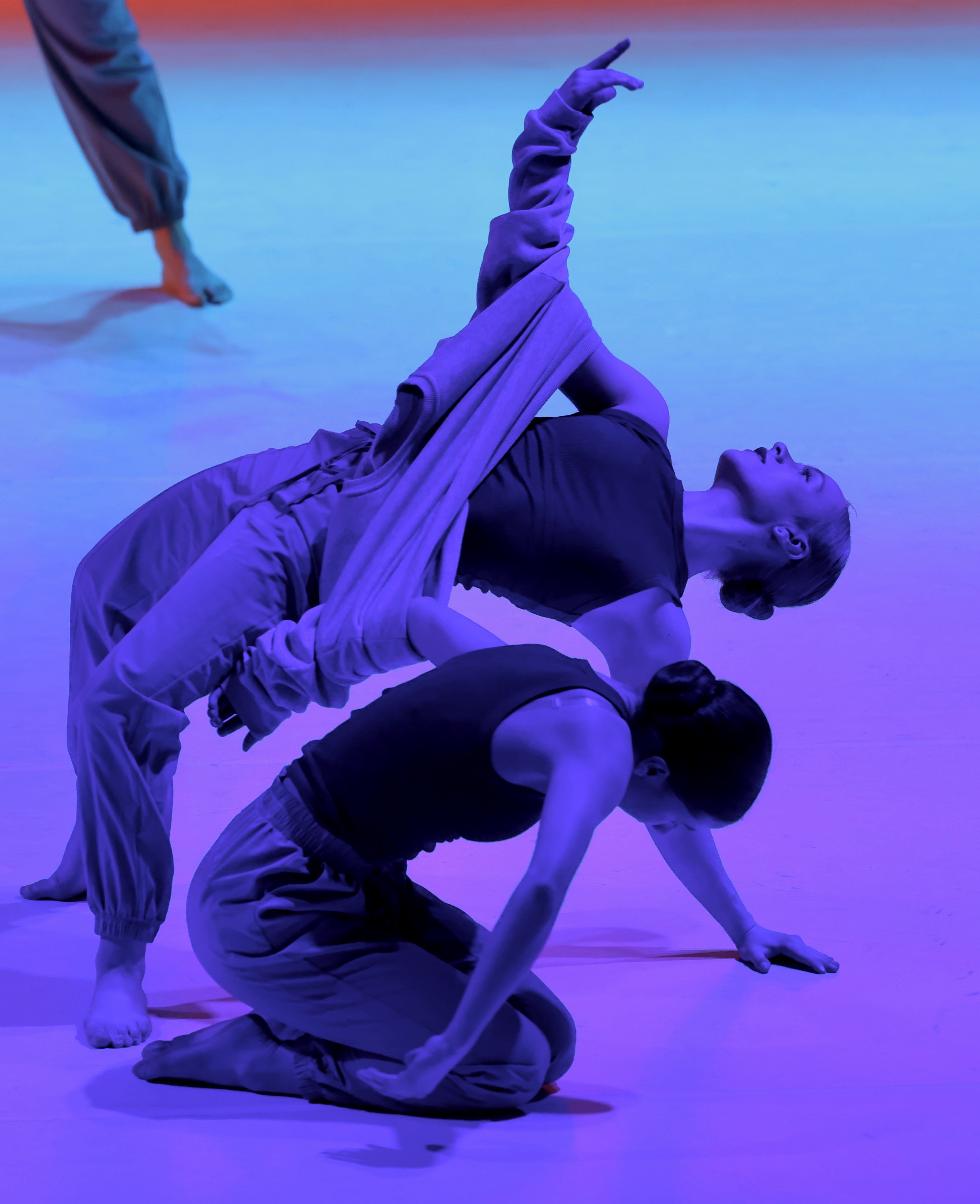 Three dancers perform on stage with colorful lighting, one lying on back with arms raised, others kneeling and leaning.