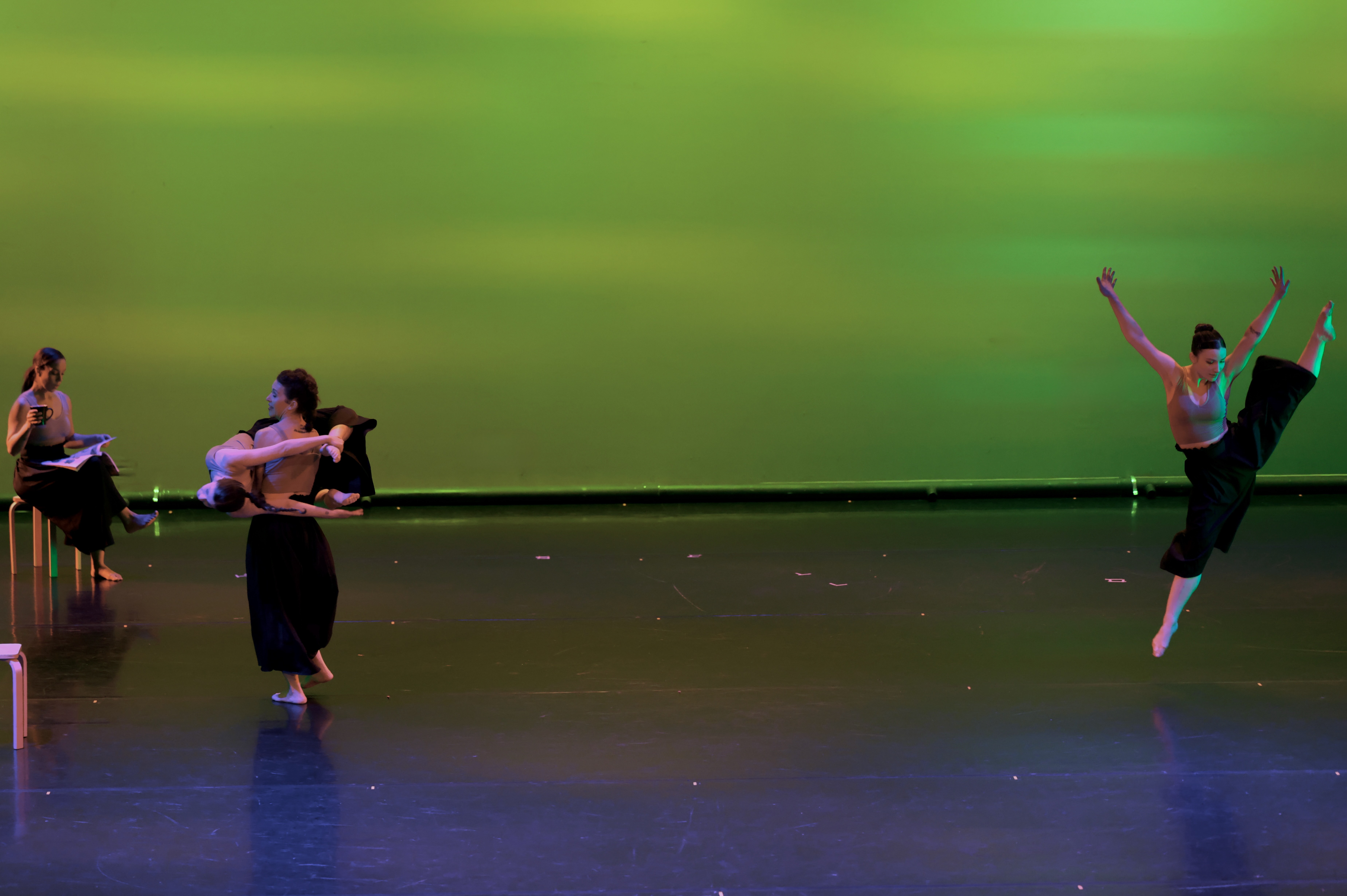Four ballet dancers perform on stage with a green background, two holding a long stick, one jumping, one sitting.