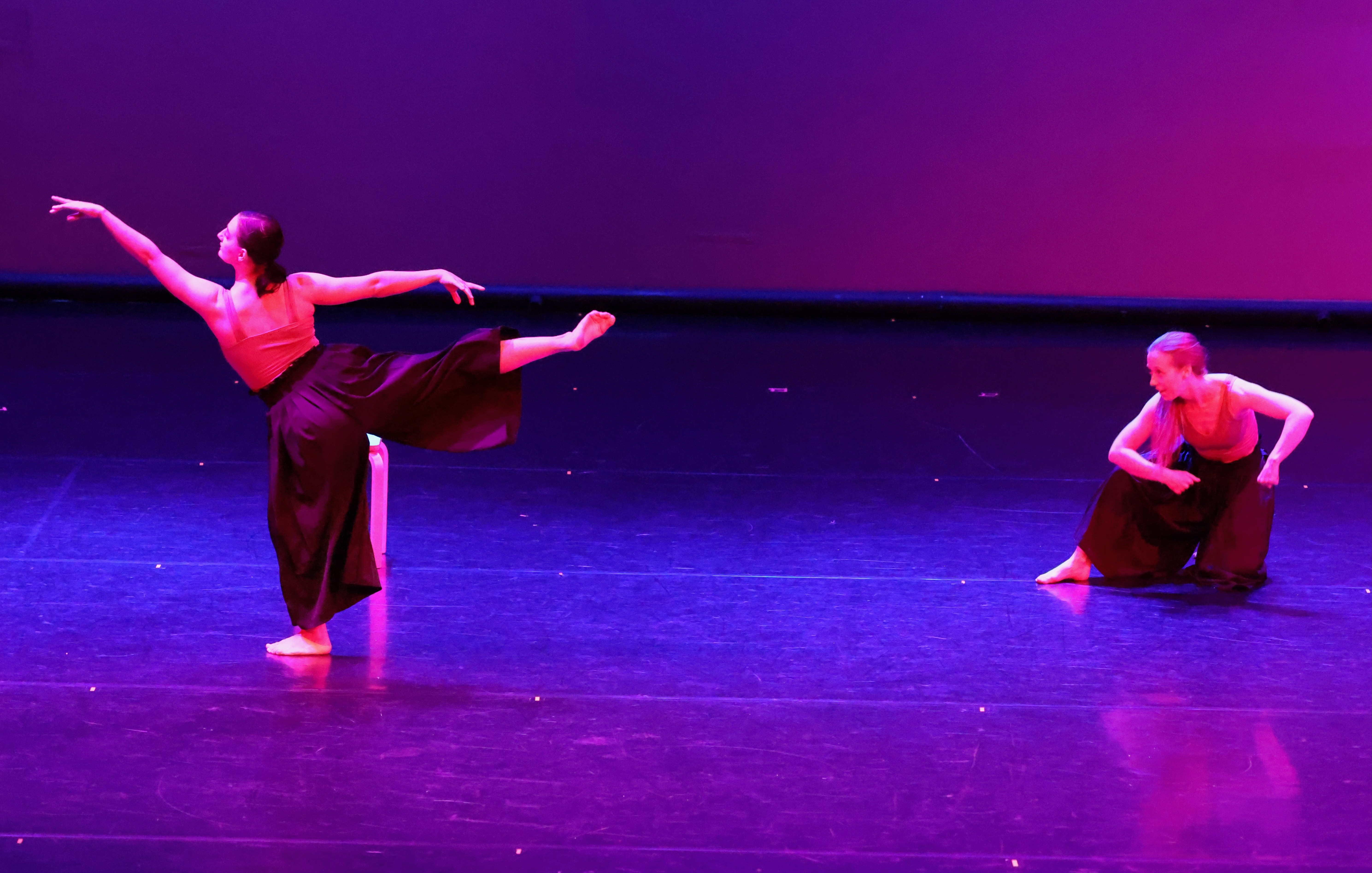 Two dancers perform on stage with purple and pink lighting, one standing and the other kneeling, both in dance poses.