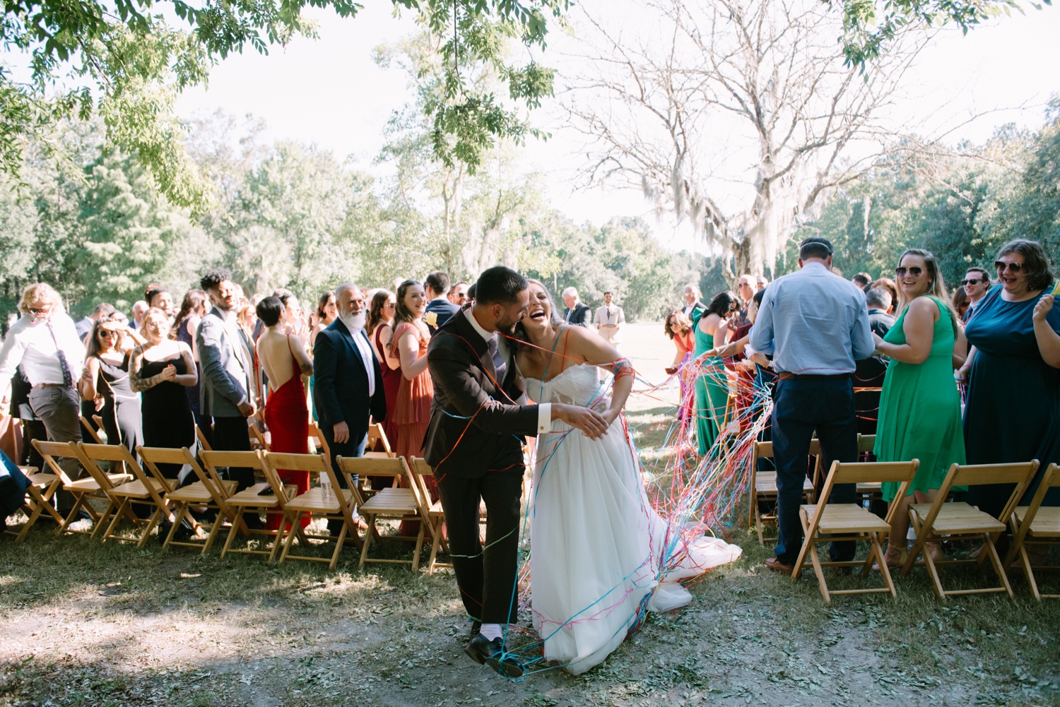 Couple dancing at outdoor wedding ceremony with guests seated on wooden chairs and trees in background.