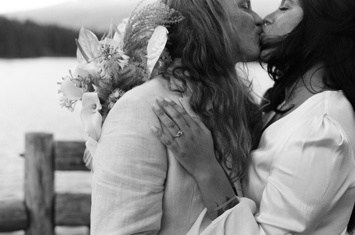 Two women kissing outdoors, one holding a bouquet, with a body of water and trees in the background.