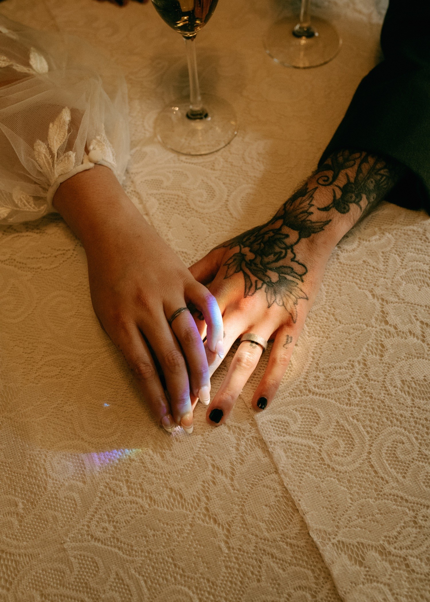 Two hands, one with a tattoo, touch on a table with two glasses in the background.