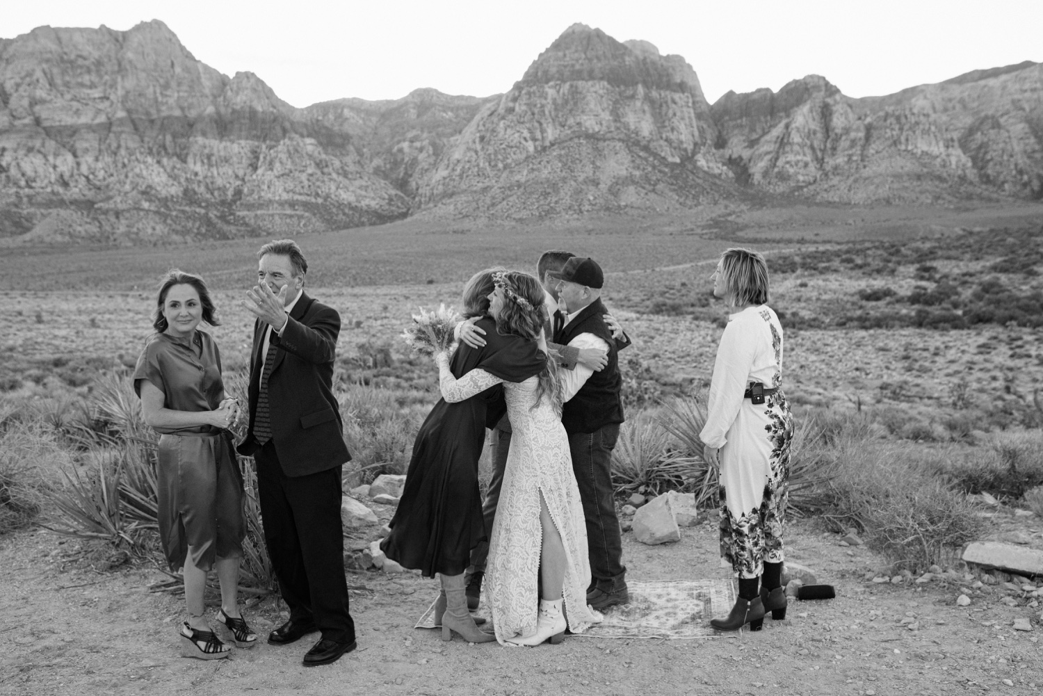 Group of six people hugging and standing outdoors with mountains in background, some smiling and others observing.