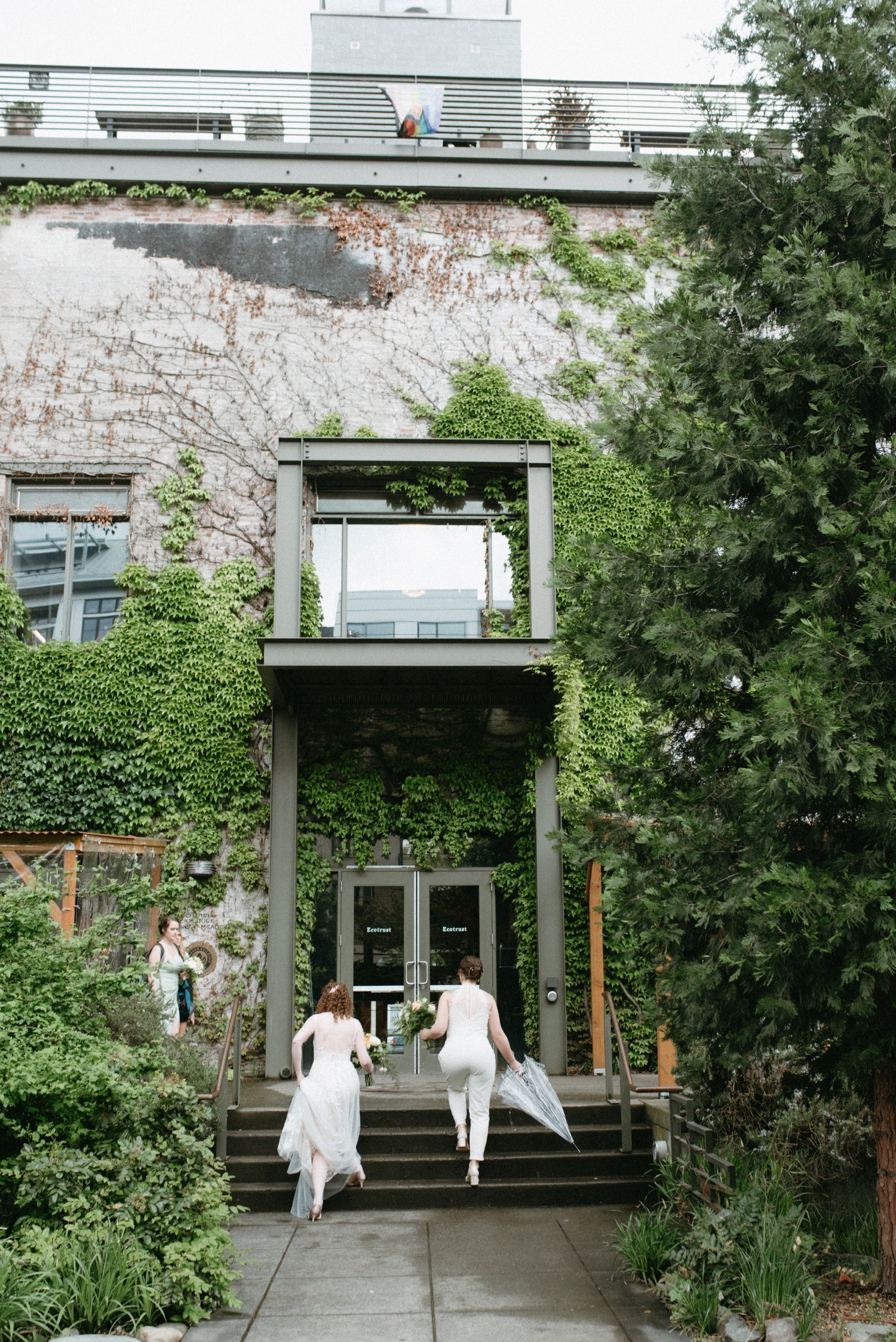 Three people walk up stairs outside a building with large glass doors, surrounded by greenery and trees.