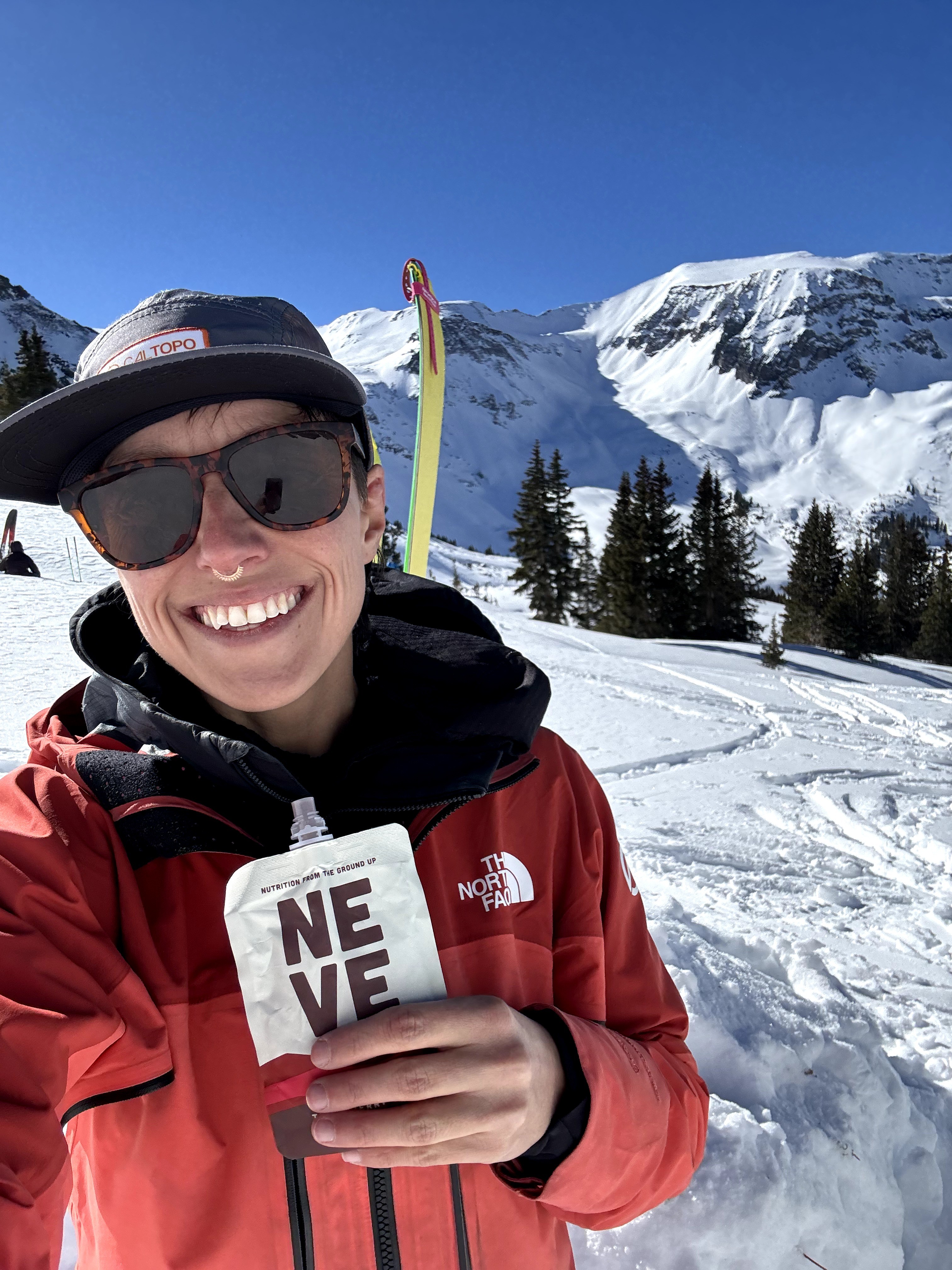 Smiling person in red jacket and sunglasses holding a ski pole in snowy mountain landscape.