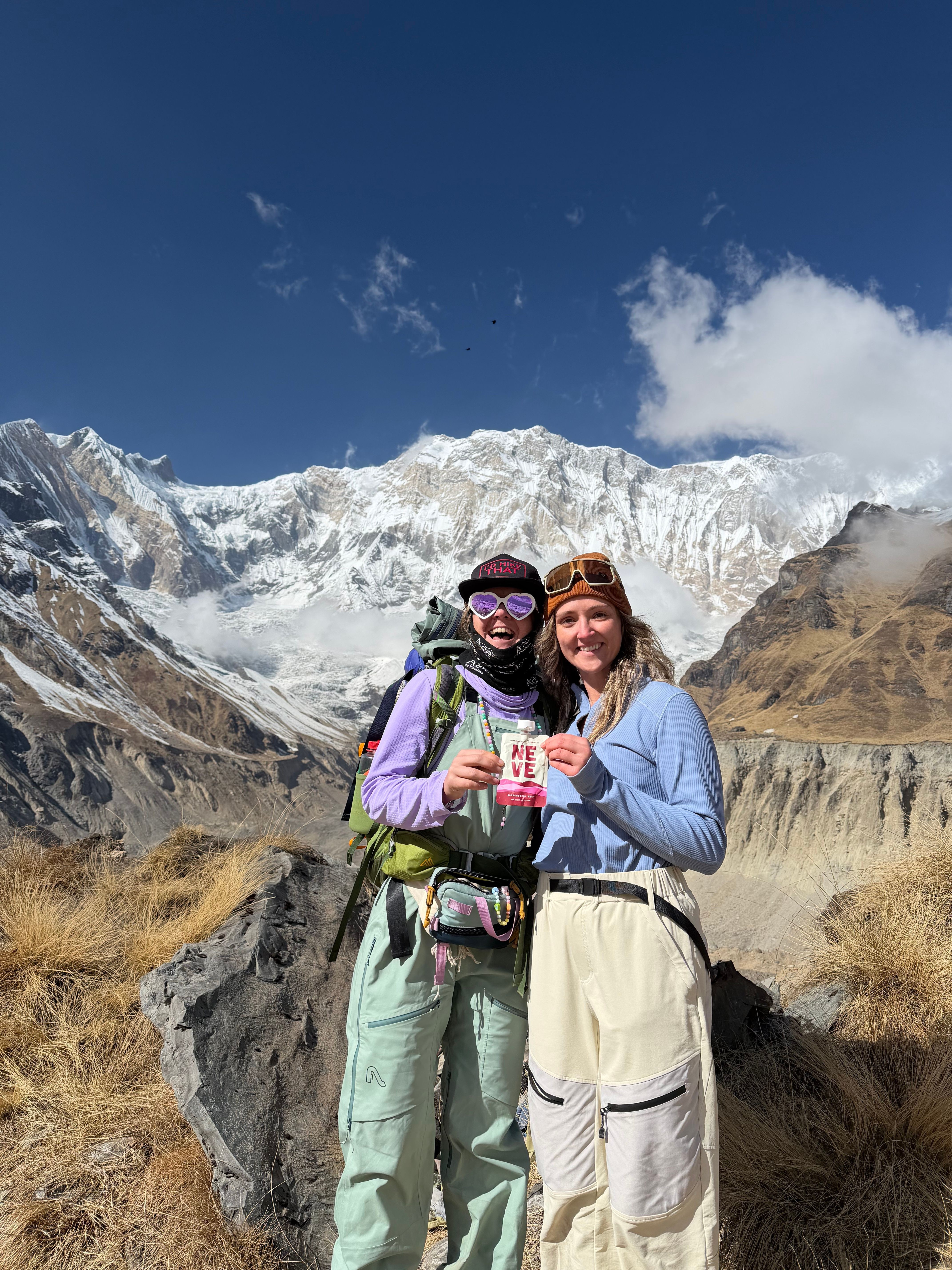 Two people stand outdoors in front of snow-capped mountains, smiling and holding a small object together.