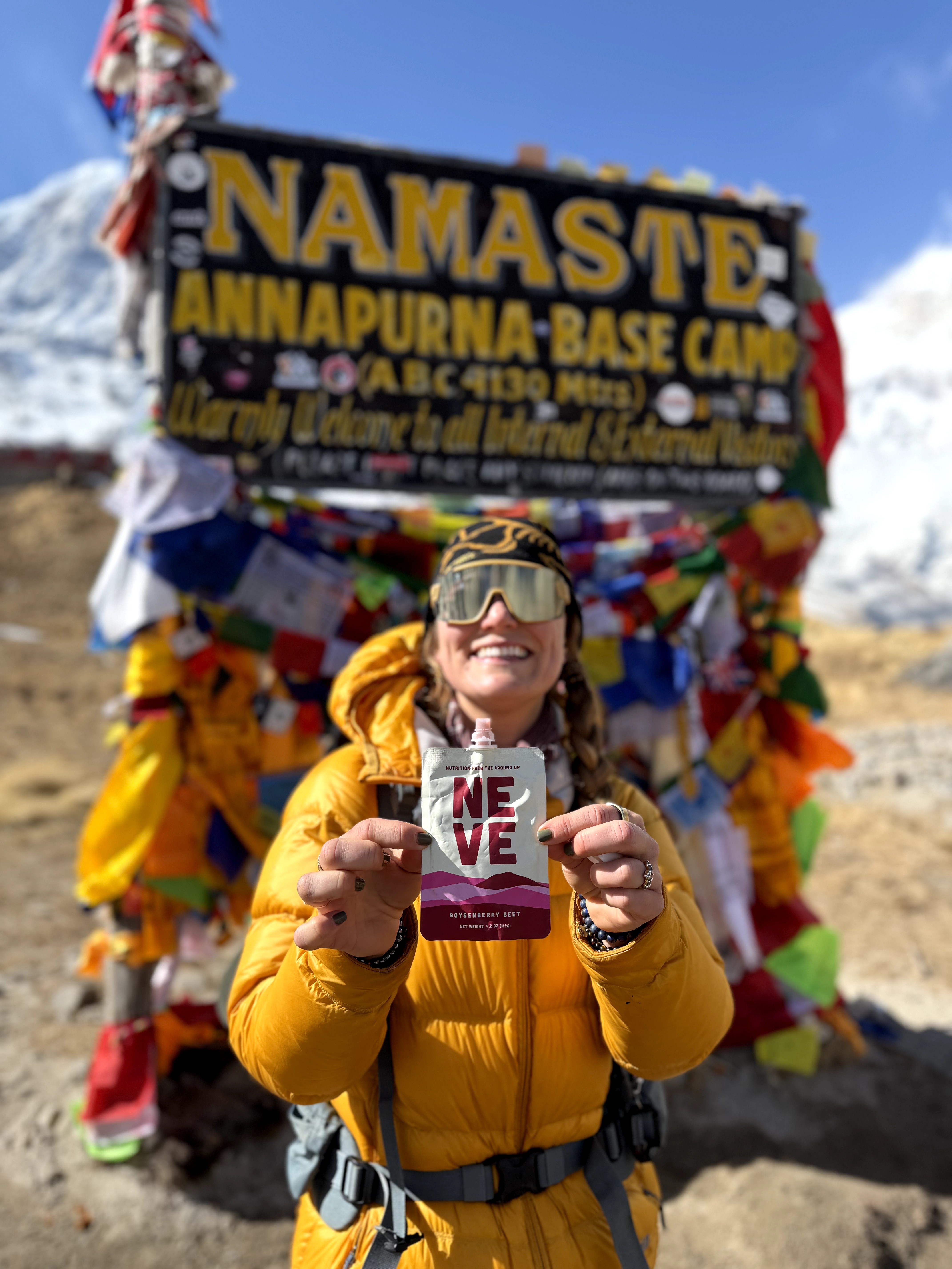 Person in yellow jacket holding a card with 'LOVE' written on it, smiling, in front of a decorated sign at Annapurna Base Camp.