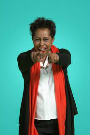 Woman smiling and pointing forward with both hands, wearing a black jacket and red scarf, against a teal background.
