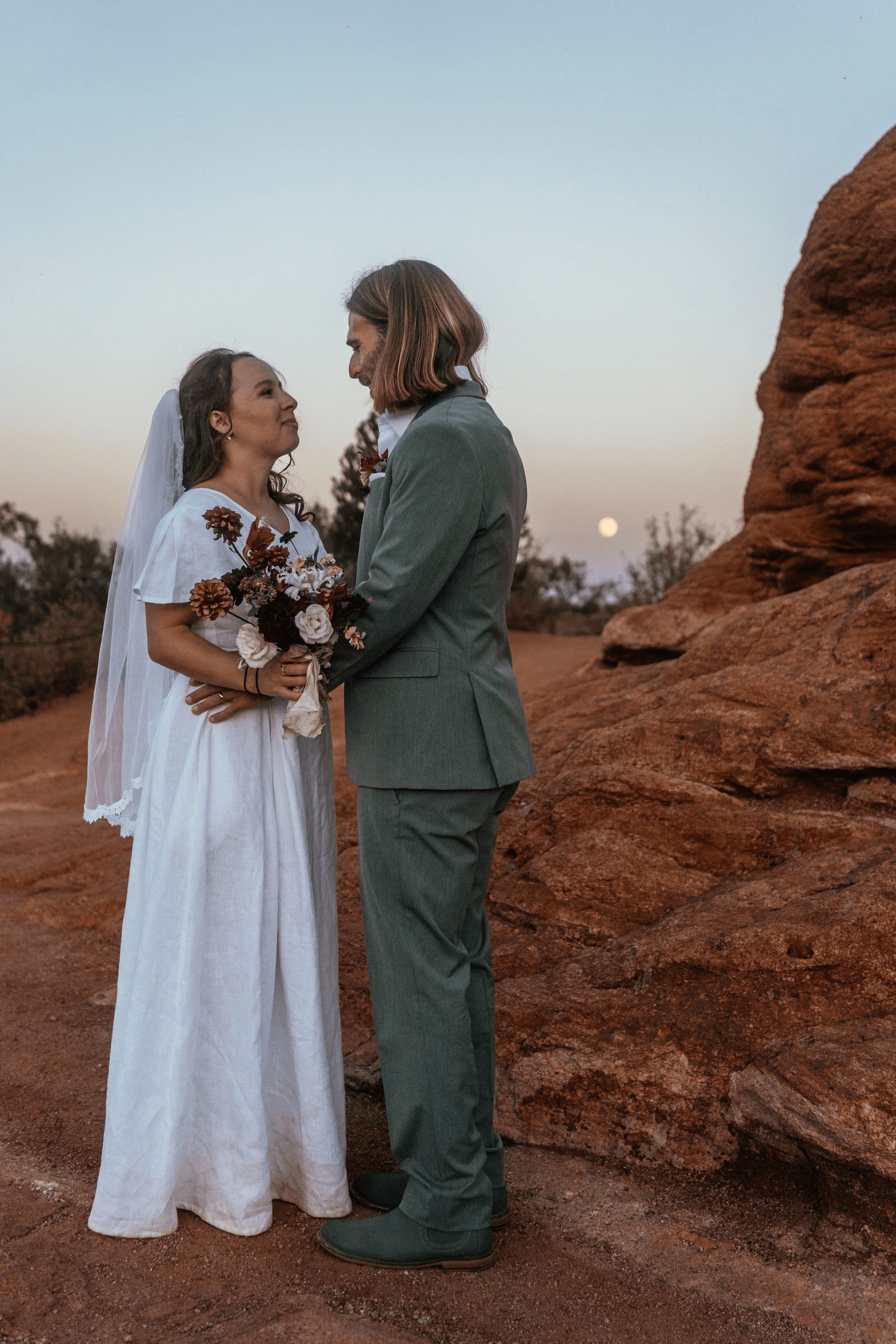 Bride and groom stand facing each other outdoors, holding hands, with a rocky landscape and clear sky in the background.