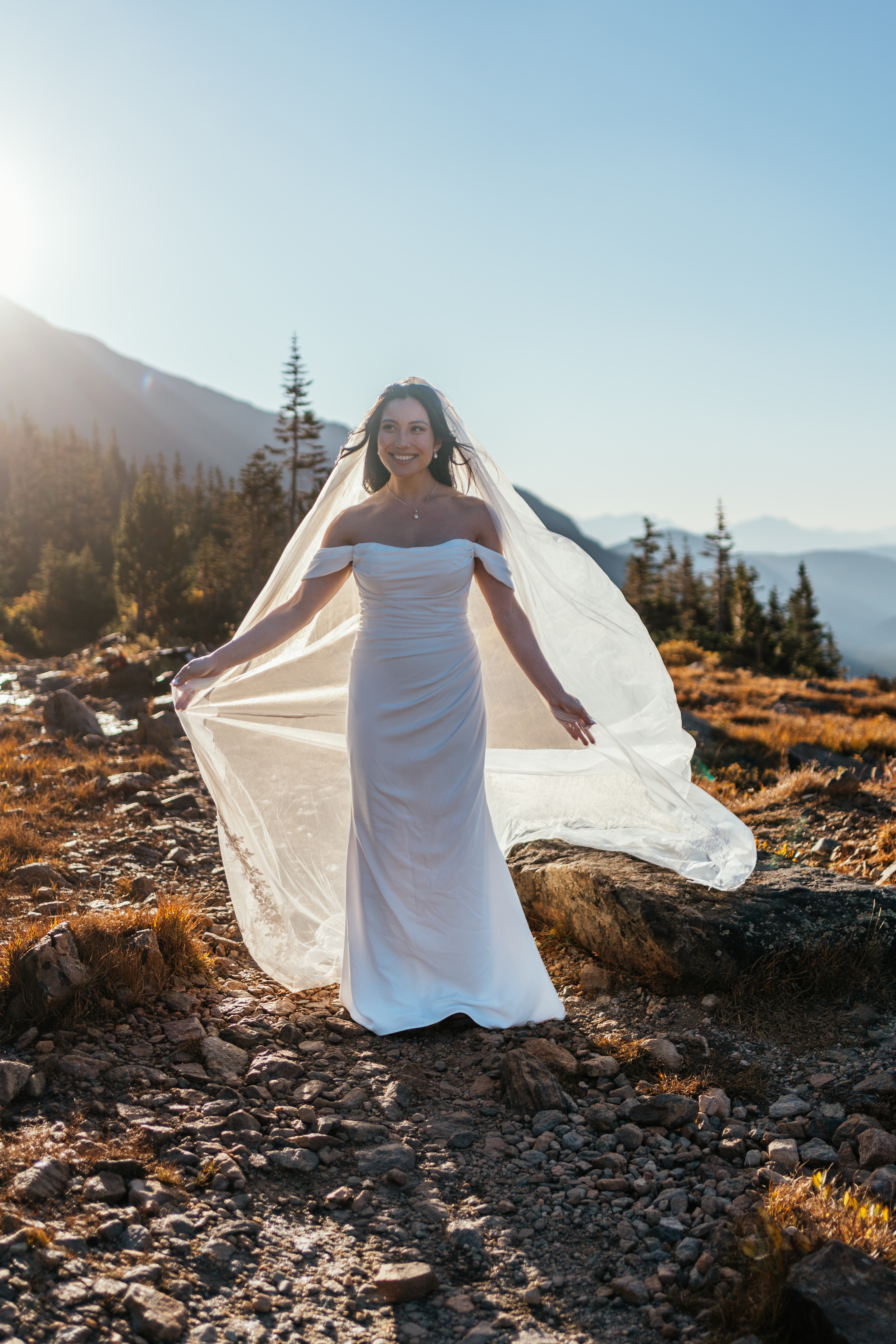 Woman in a white wedding dress standing outdoors on rocky terrain with mountains and trees in the background.