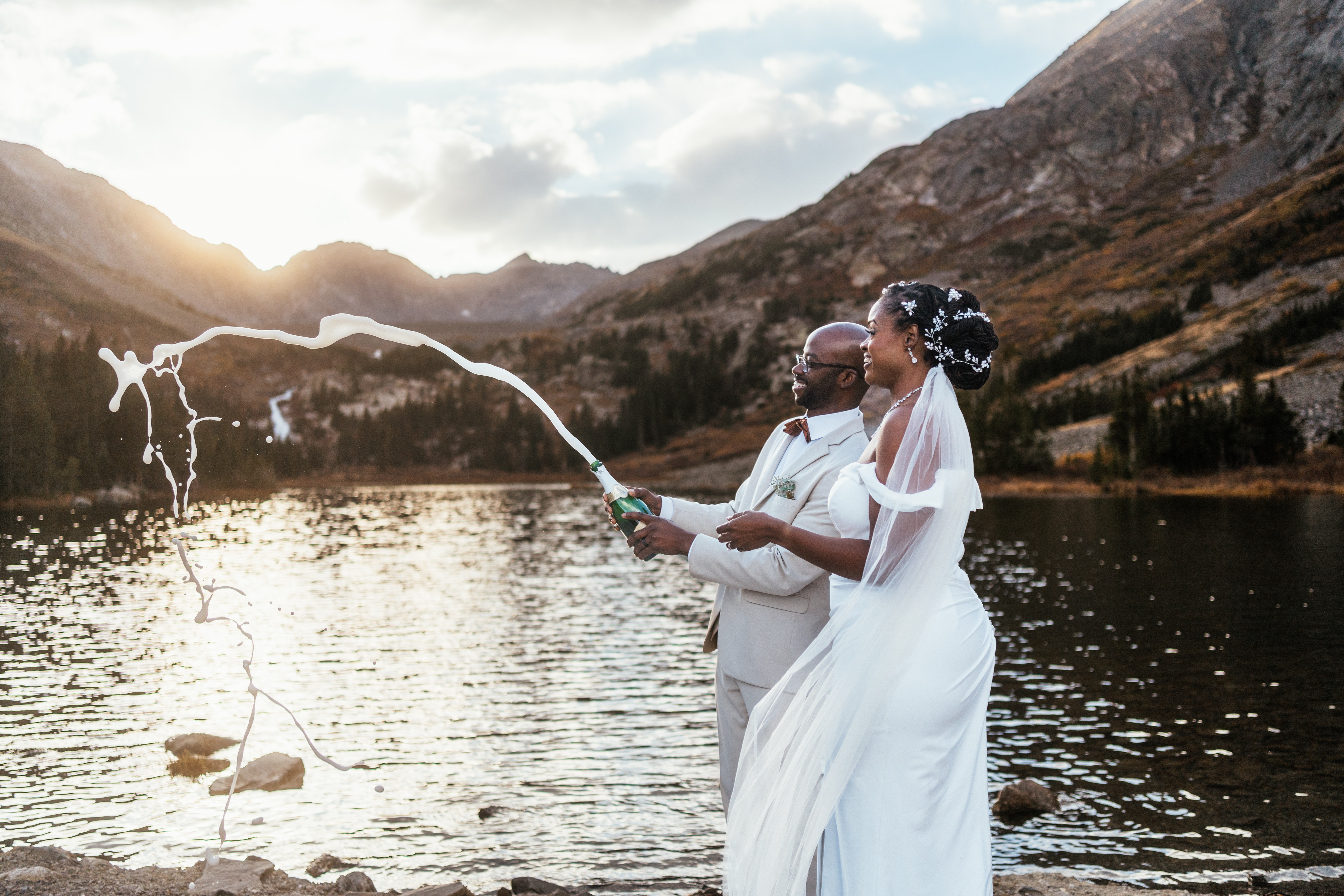 Couple in white wedding attire standing by a lake with mountains in the background, holding a champagne bottle with water splashing.