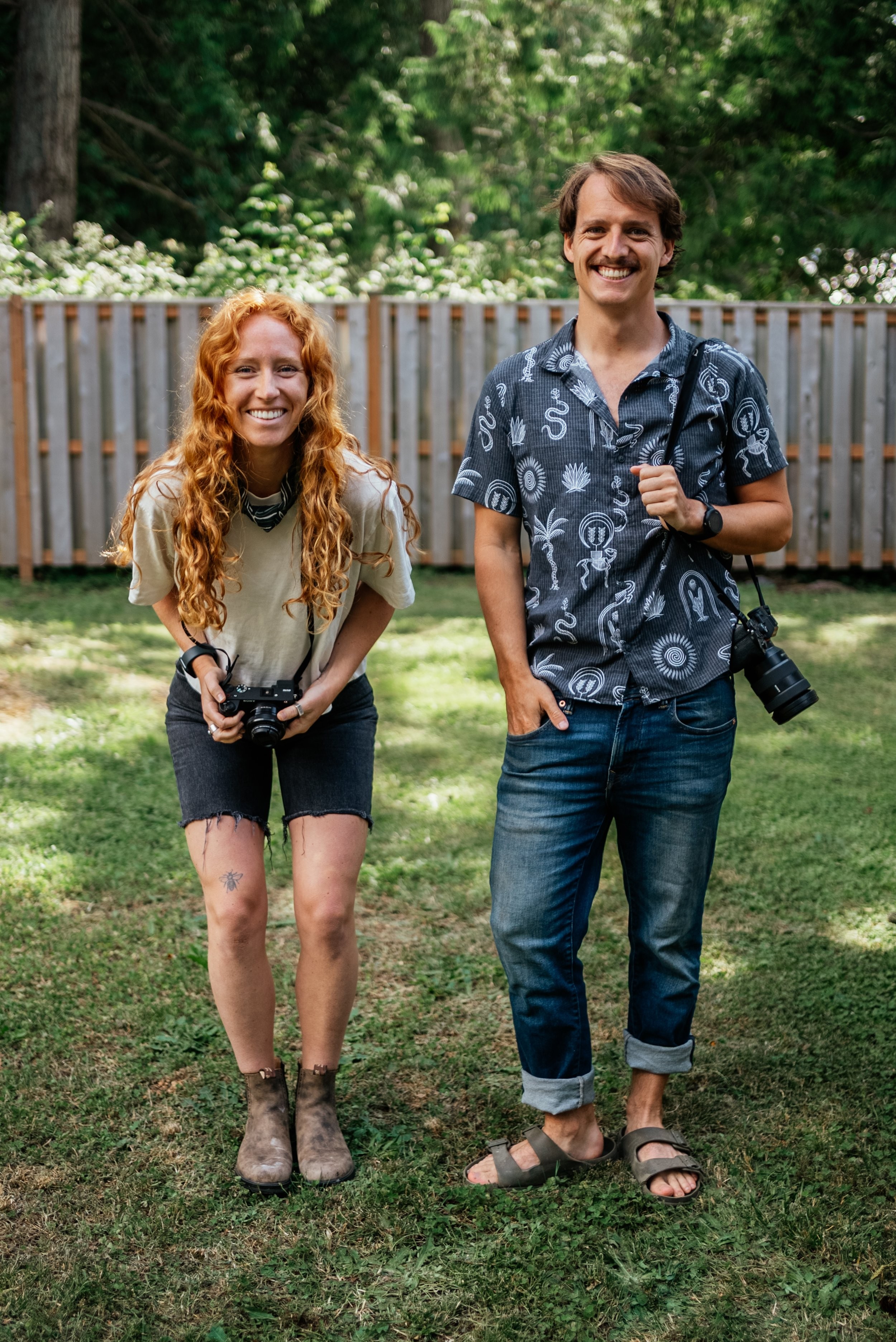 Two smiling people standing on grass in front of a wooden fence, holding cameras, outdoors with trees in background.