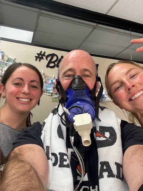 Three people smiling indoors, one wearing a mask connected to a device, background with signs and shelves.