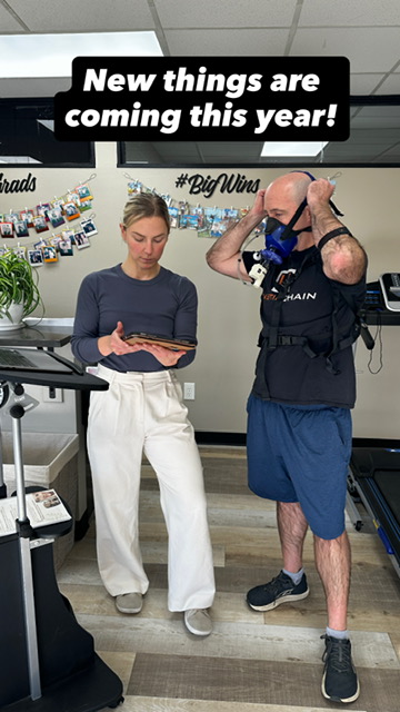 Two people standing indoors, one holding a tablet, the other adjusting a camera on his head, with a sign above them.