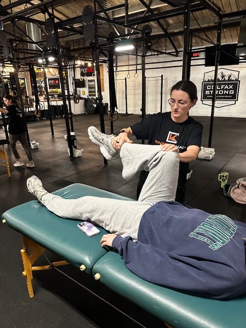 Person lying on a massage table receiving a leg stretch from a therapist in a gym.