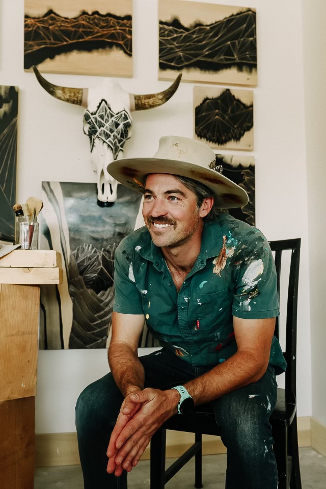 Man wearing a hat and patterned shirt sitting on a chair, smiling, with artwork and a skull mounted on the wall behind him.