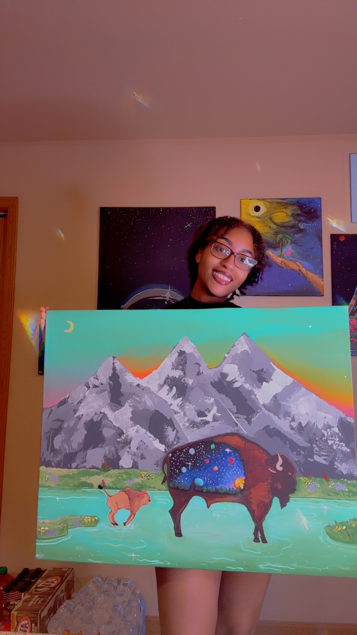 Person holding colorful painting of mountains, animals, and a rainbow, smiling indoors.