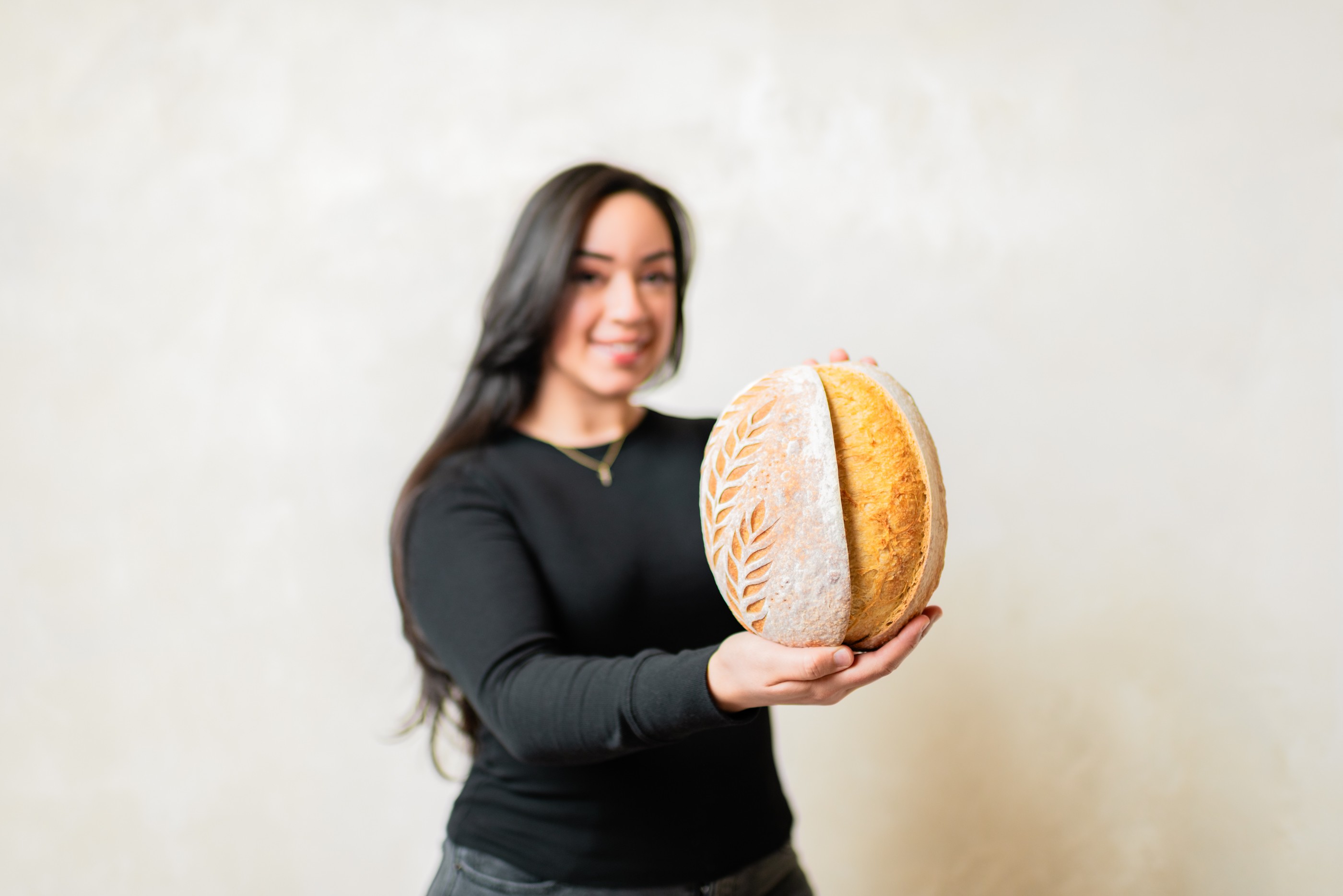 Woman with long dark hair smiling holding a round bread with a patterned crust.