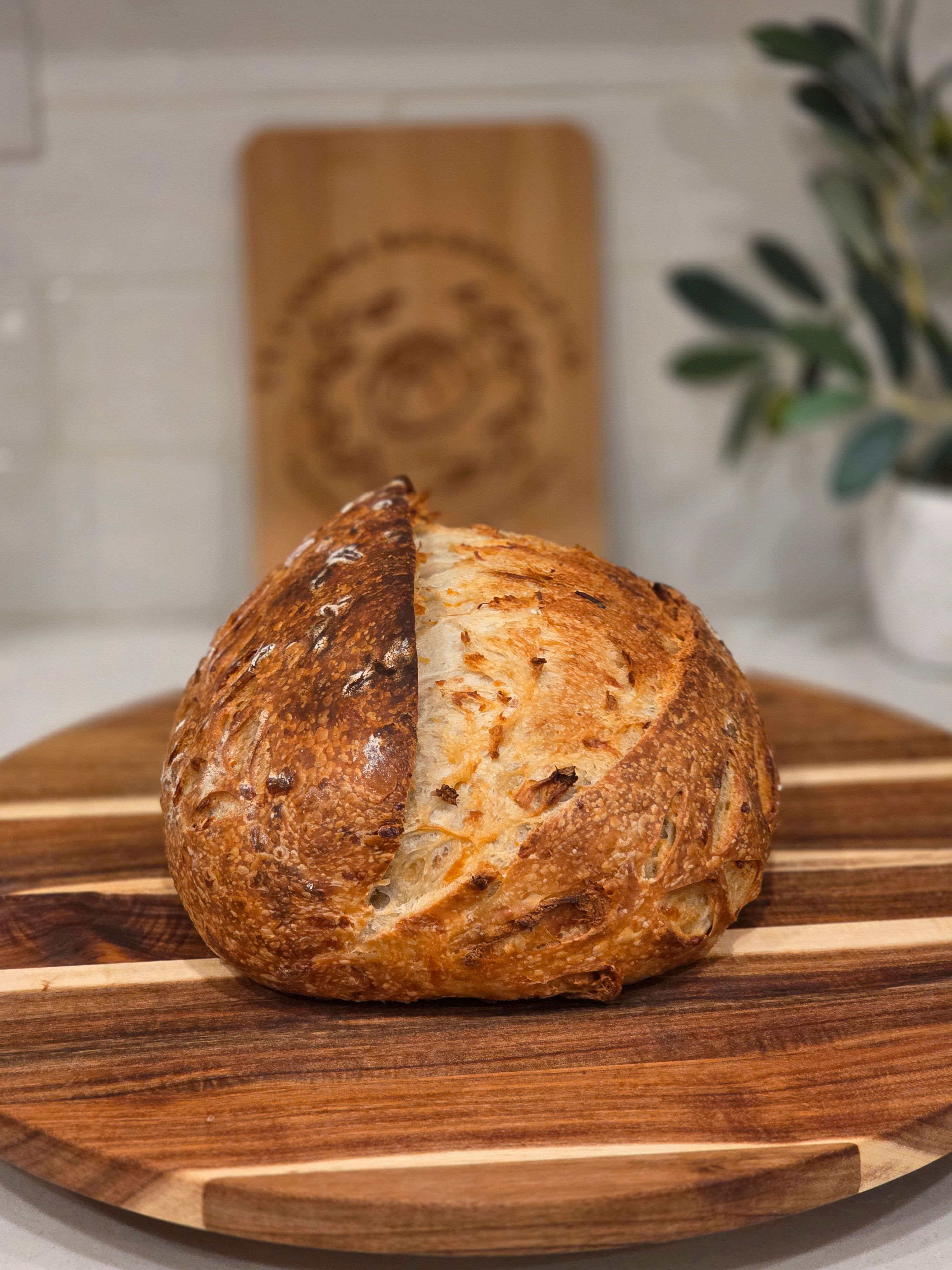 Round bread loaf with a crusty surface on a wooden cutting board, with a blurred background including a plant and a wooden sign.