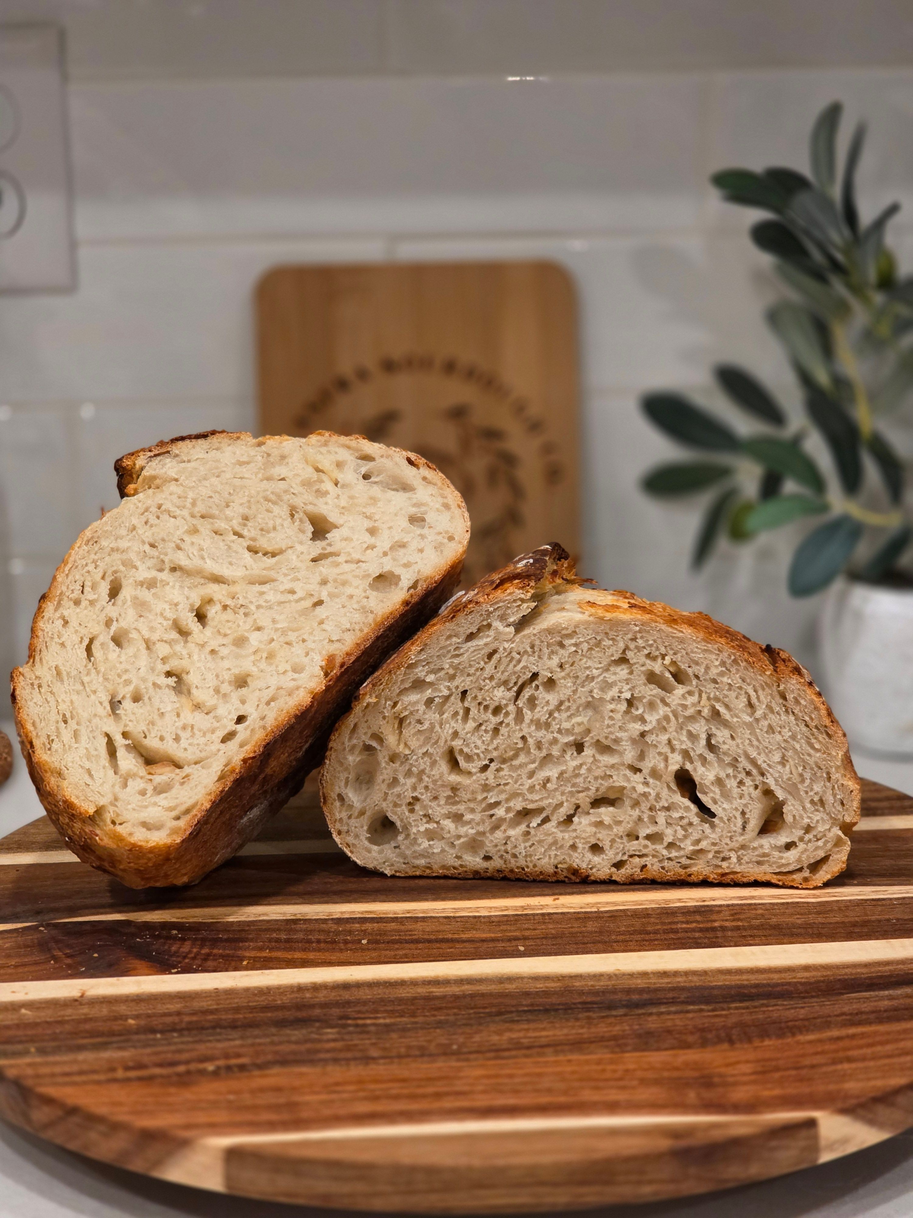 Two halves of a loaf of bread on a wooden cutting board, with a plant and a cutting board in the background.