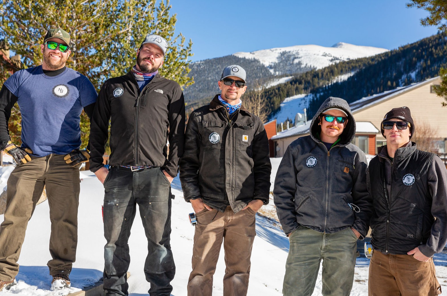 Six people standing outdoors in snowy landscape with mountains and trees in background.