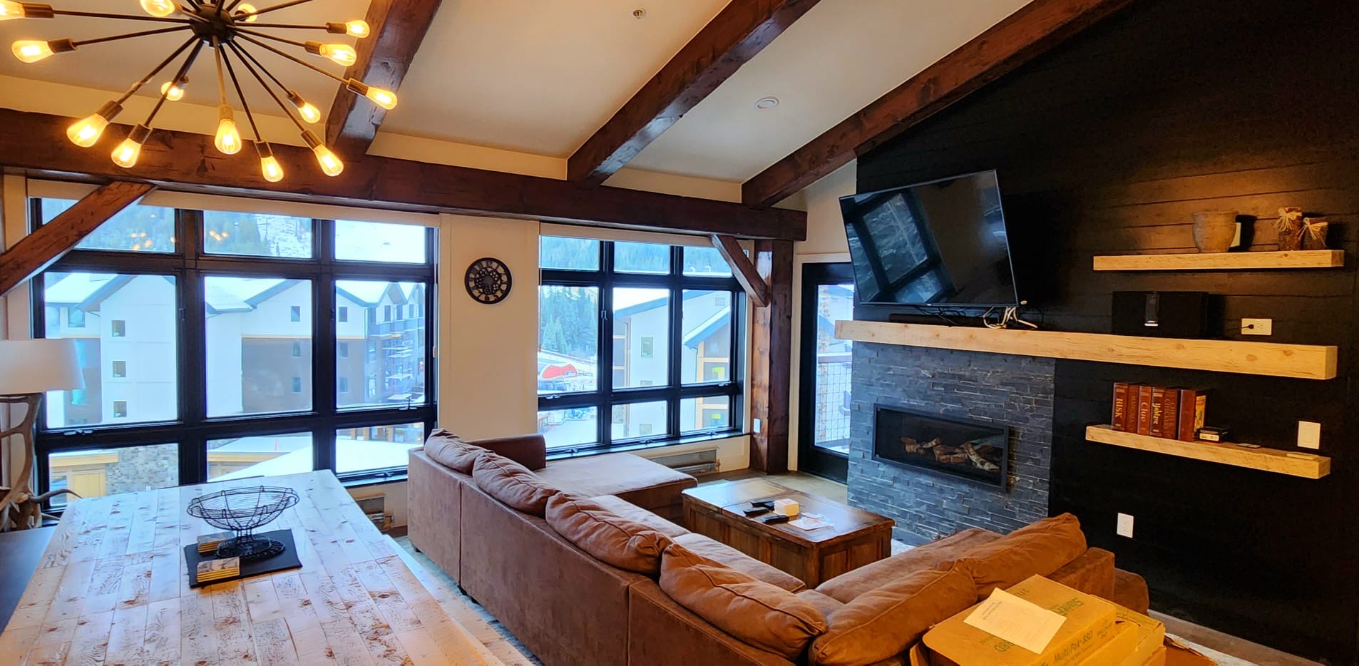 Living room with large windows, brown sofa, fireplace, mounted TV, and wooden ceiling beams.