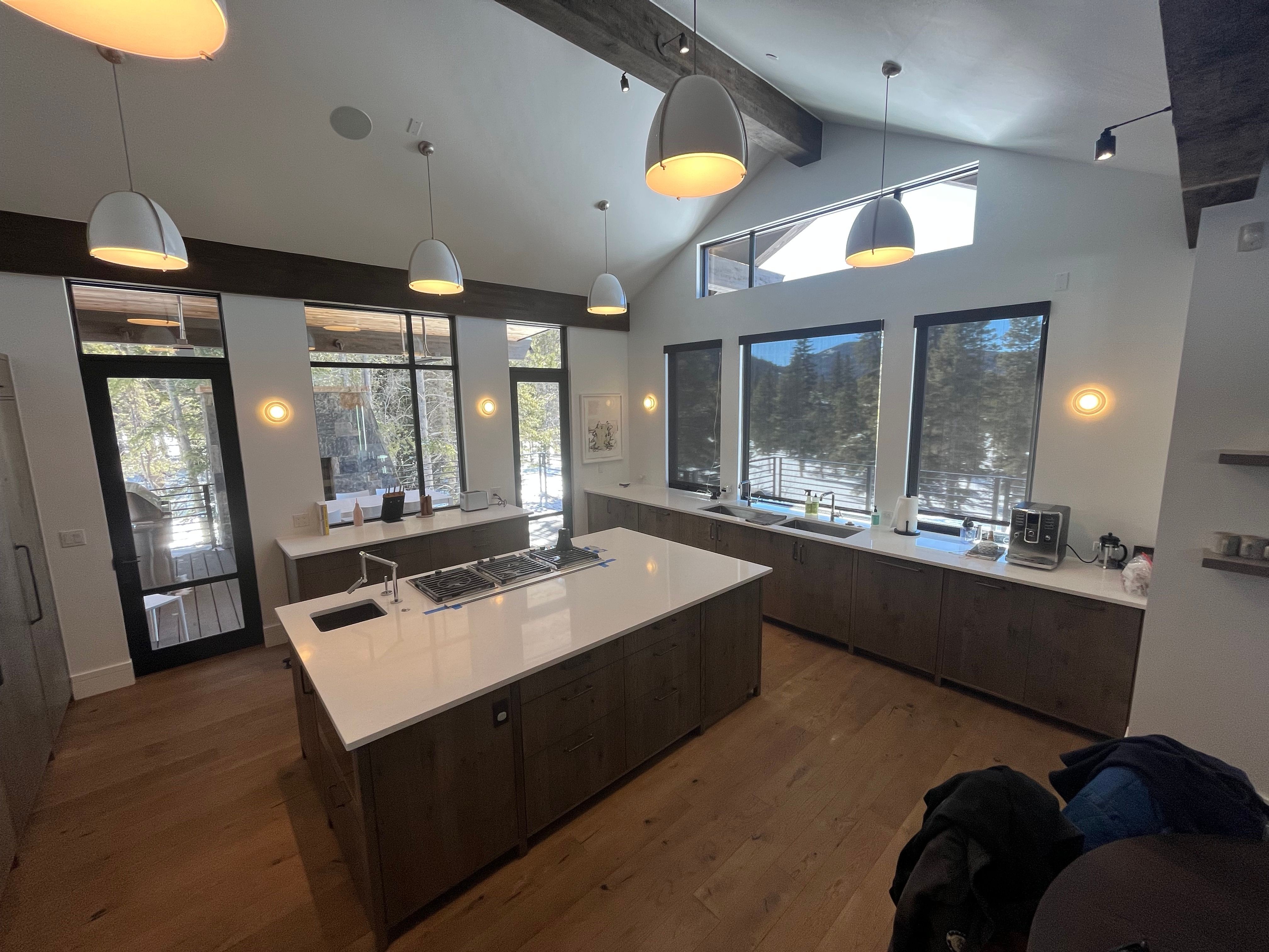 Modern kitchen with large island, multiple windows, pendant lights, and a view outside. Wooden floor and cabinets.