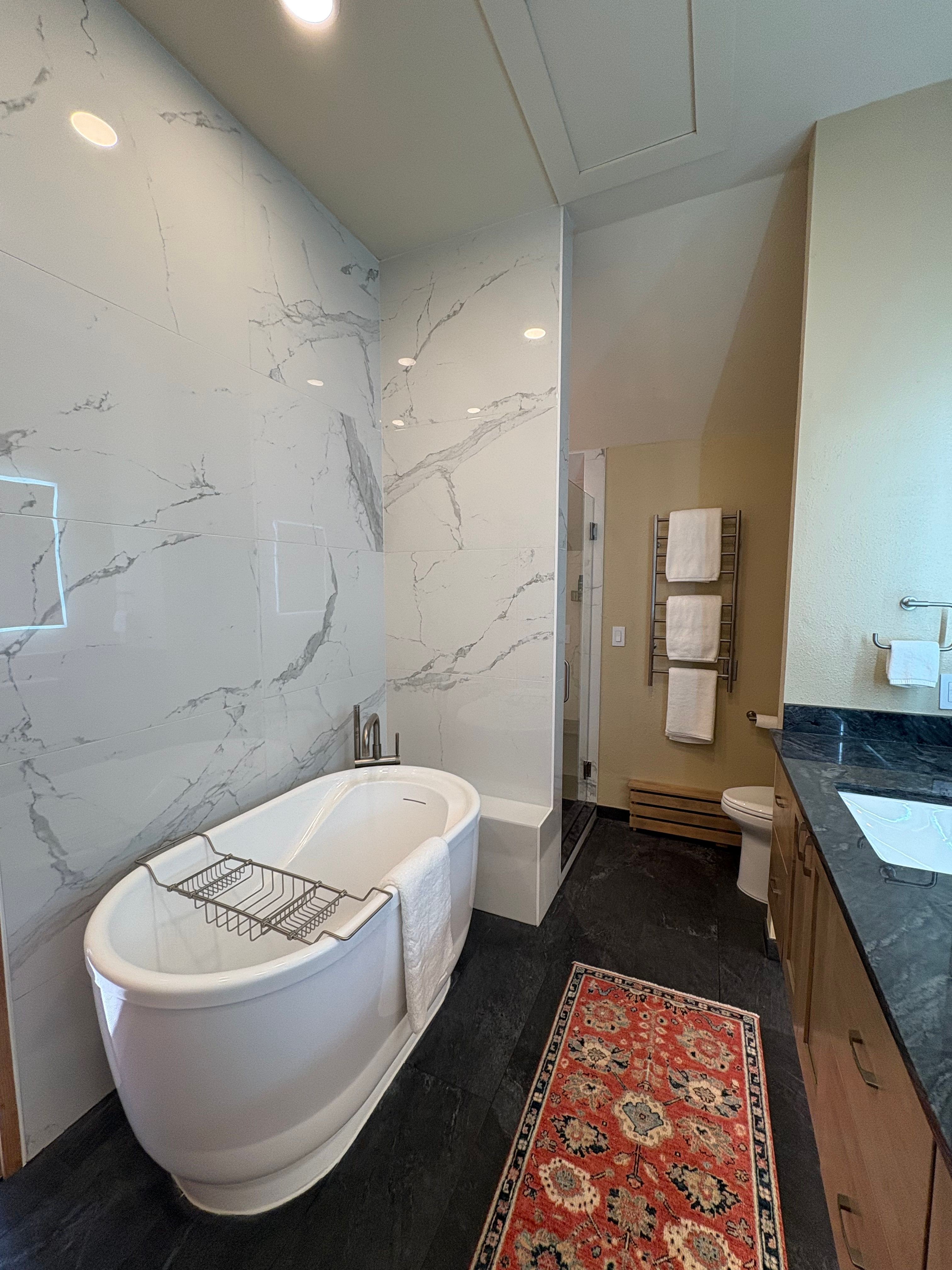 Bathroom with white marble wall, modern bathtub, black floor, and a colorful rug. Towel rack and countertop visible.