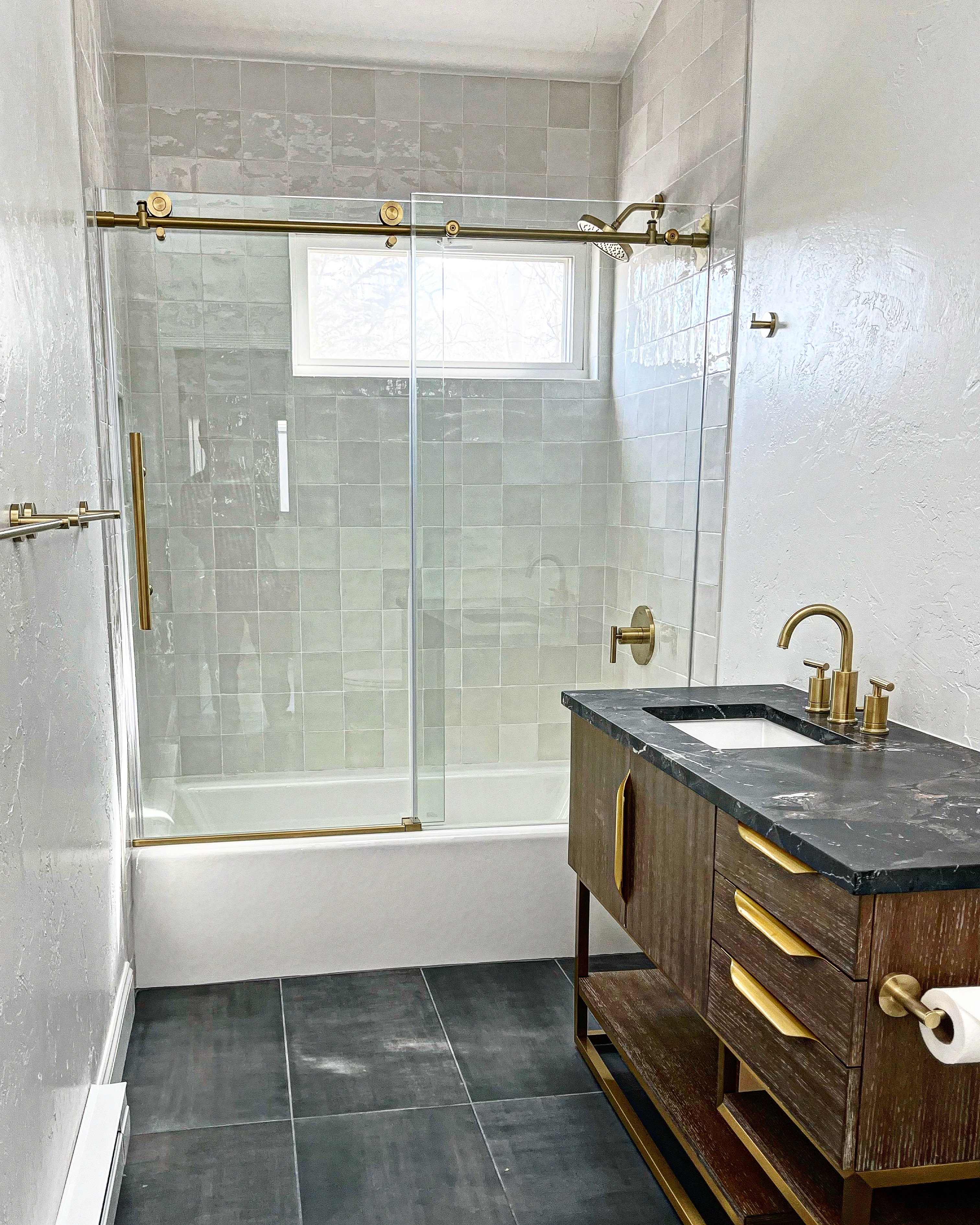 Bathroom with bathtub, glass shower doors, window, and wooden vanity with black countertop and gold fixtures.