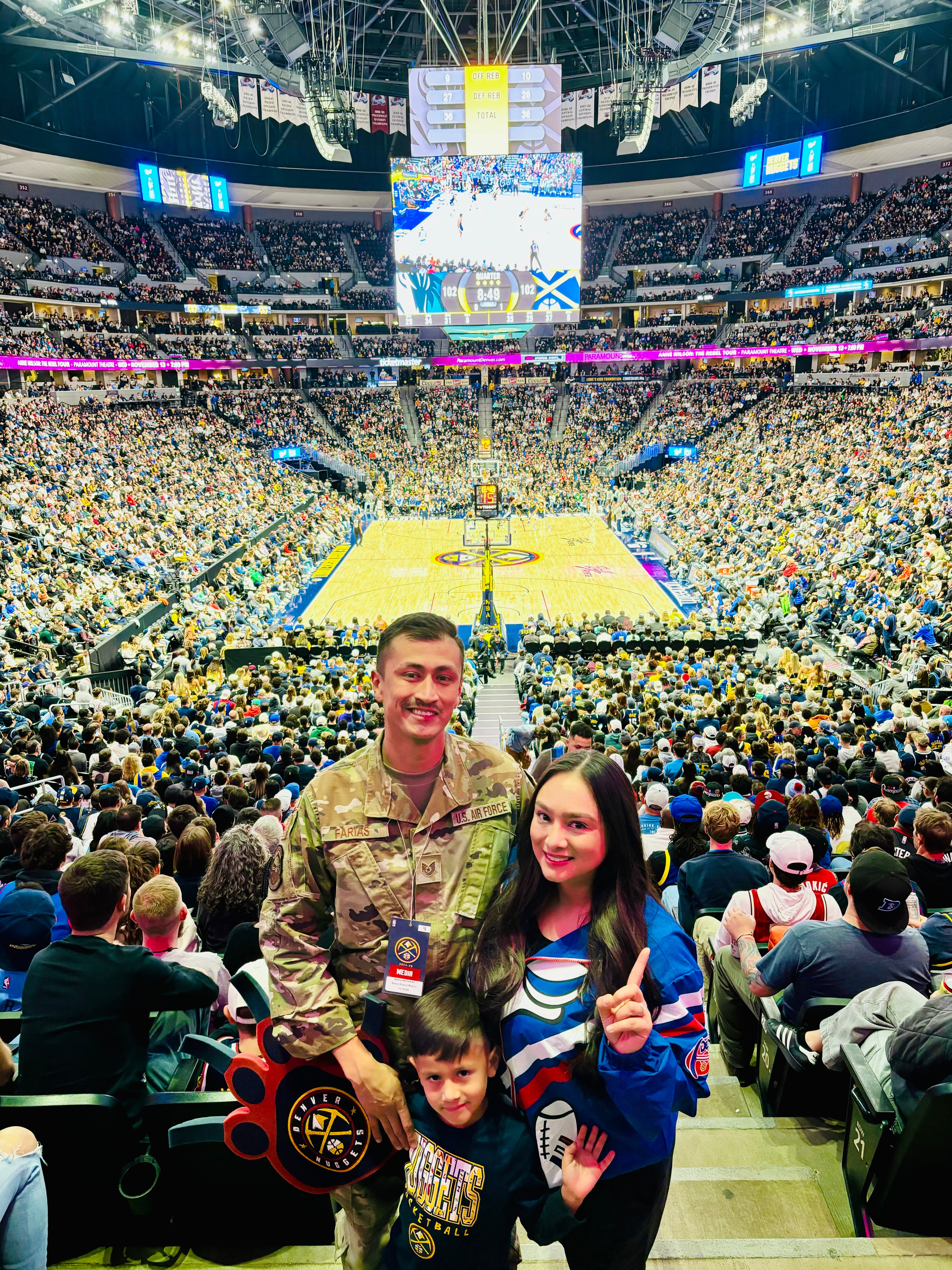 Three people, a man in military uniform, a woman, and a child, pose in front of a basketball court filled with spectators.