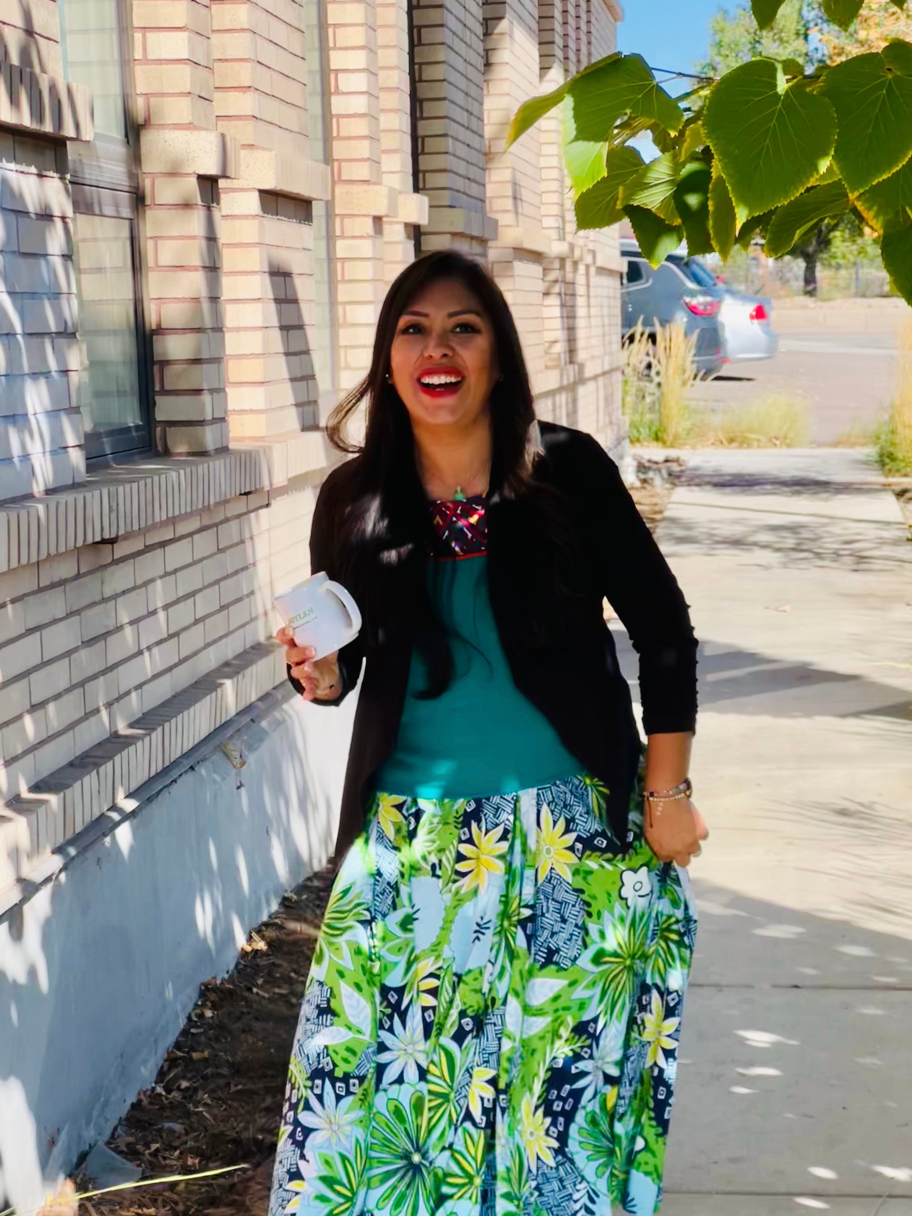 Woman with long dark hair smiling, wearing a black jacket and colorful skirt, standing outdoors near a brick building.