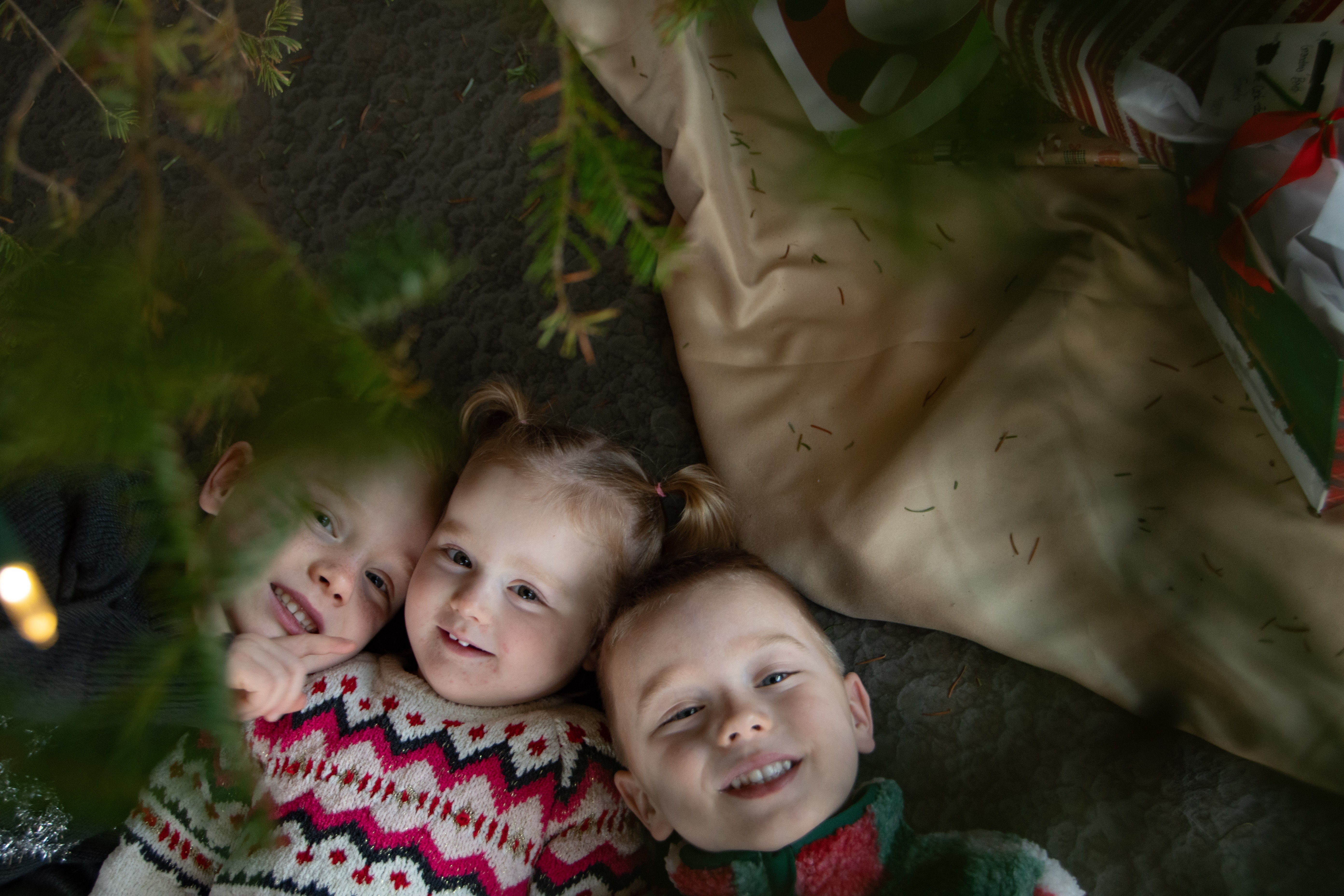 Three children lying on the ground, smiling and looking up at the camera, with greenery and a beige cloth nearby.