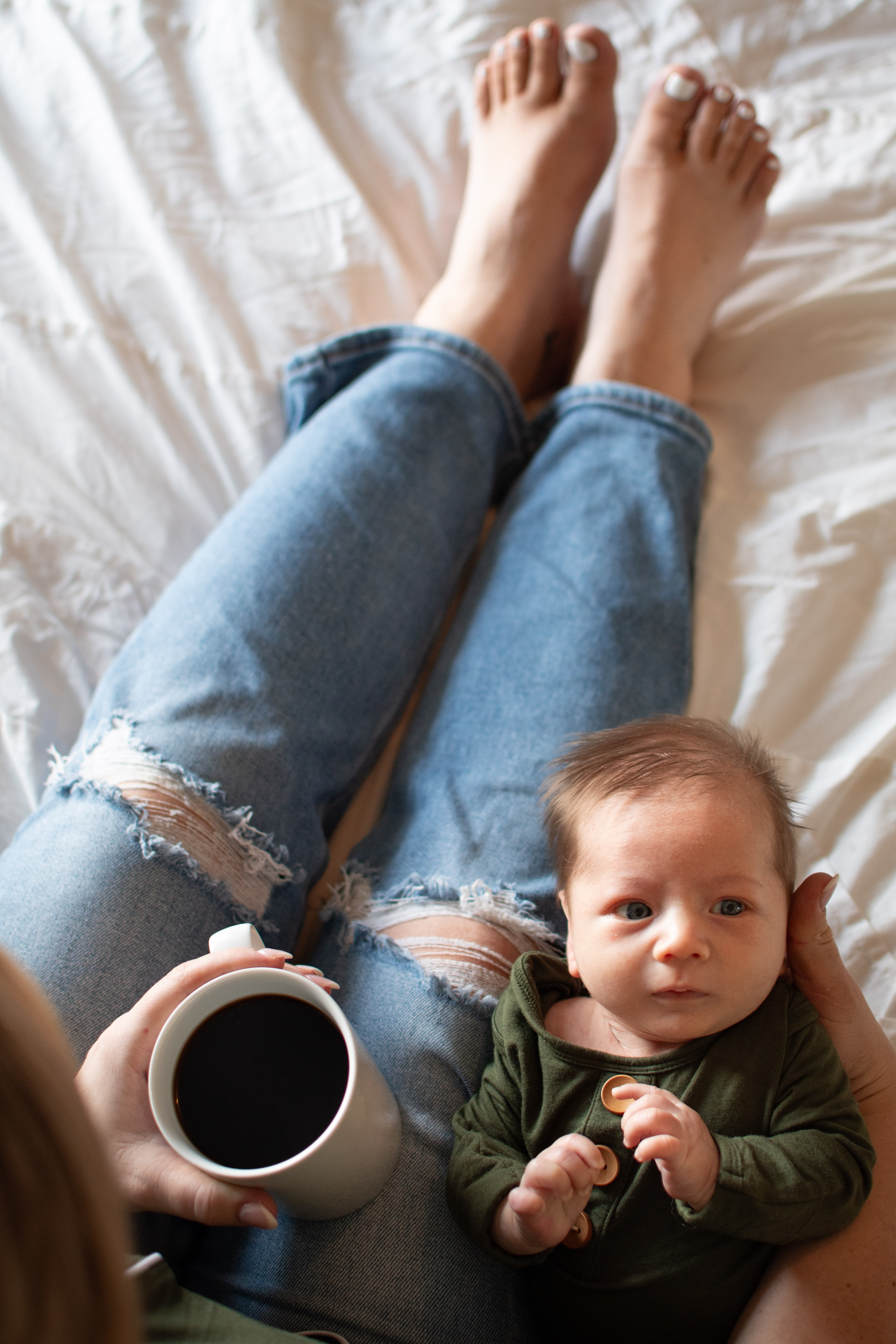 Person's legs in ripped jeans, a baby lying on bed holding a mug, and a white bedsheet background.