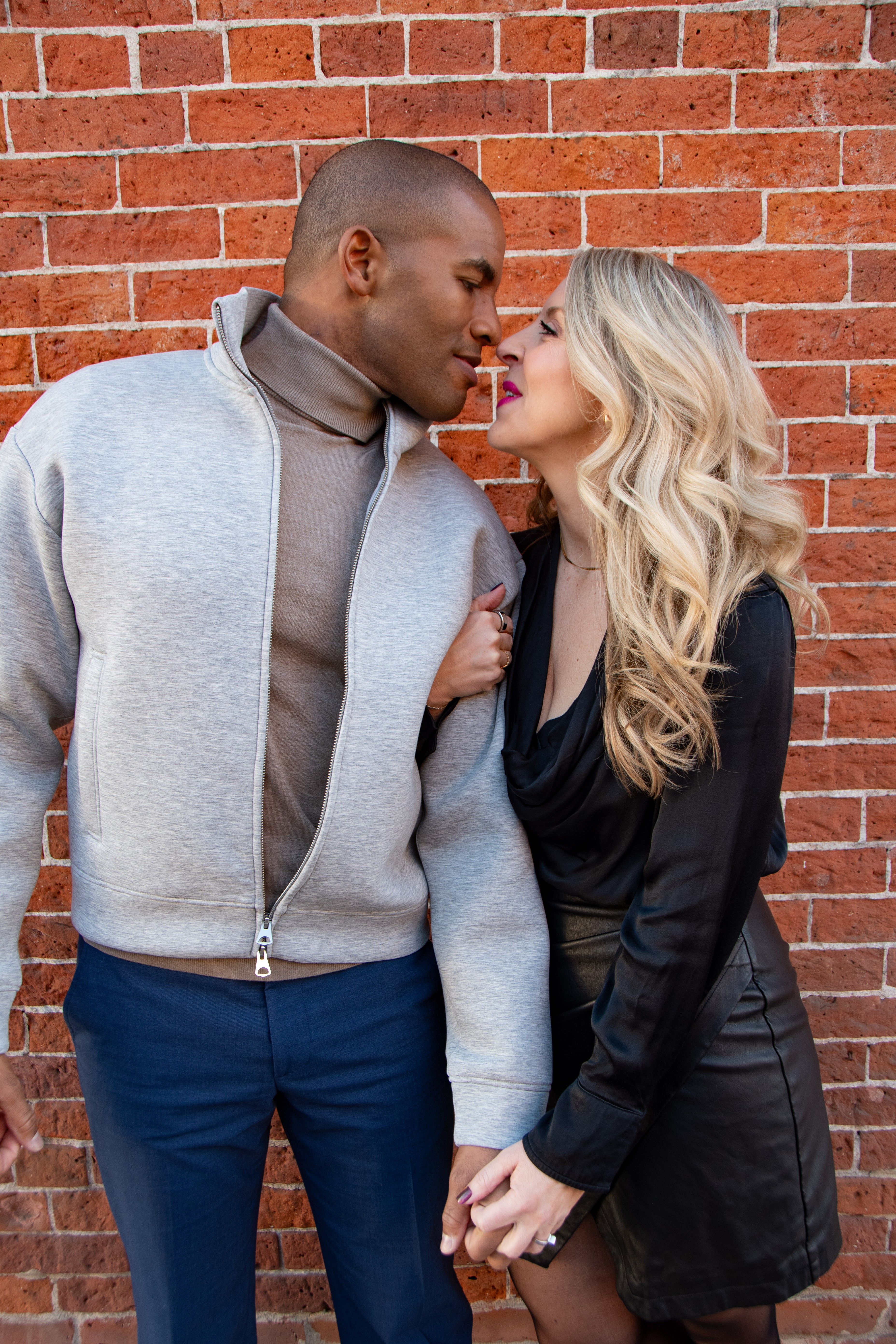 Couple standing close against a brick wall, about to kiss, holding hands, woman with blonde hair, man with short hair.