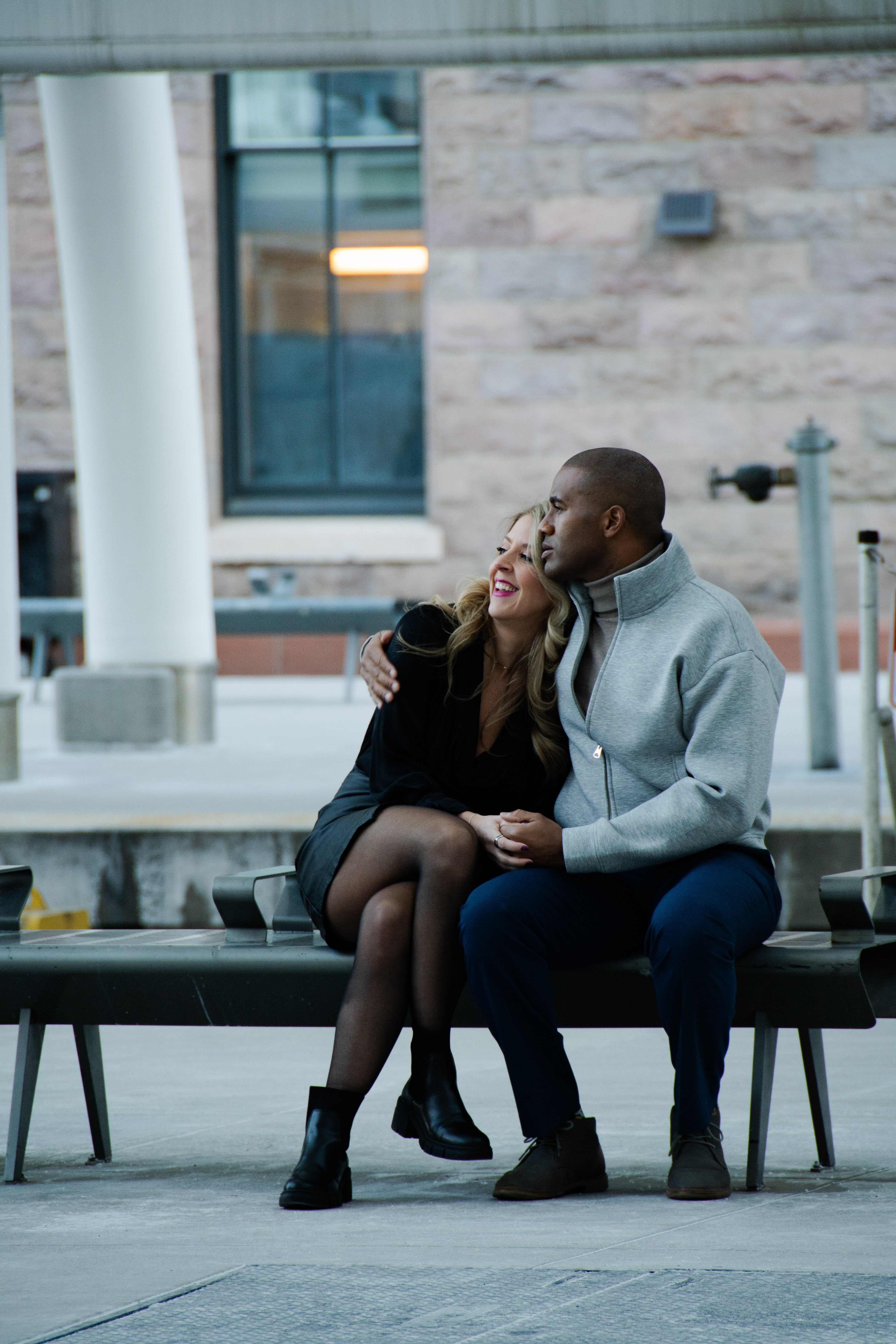A couple sitting on a bench, with the woman smiling and the man kissing her cheek, in an urban setting.