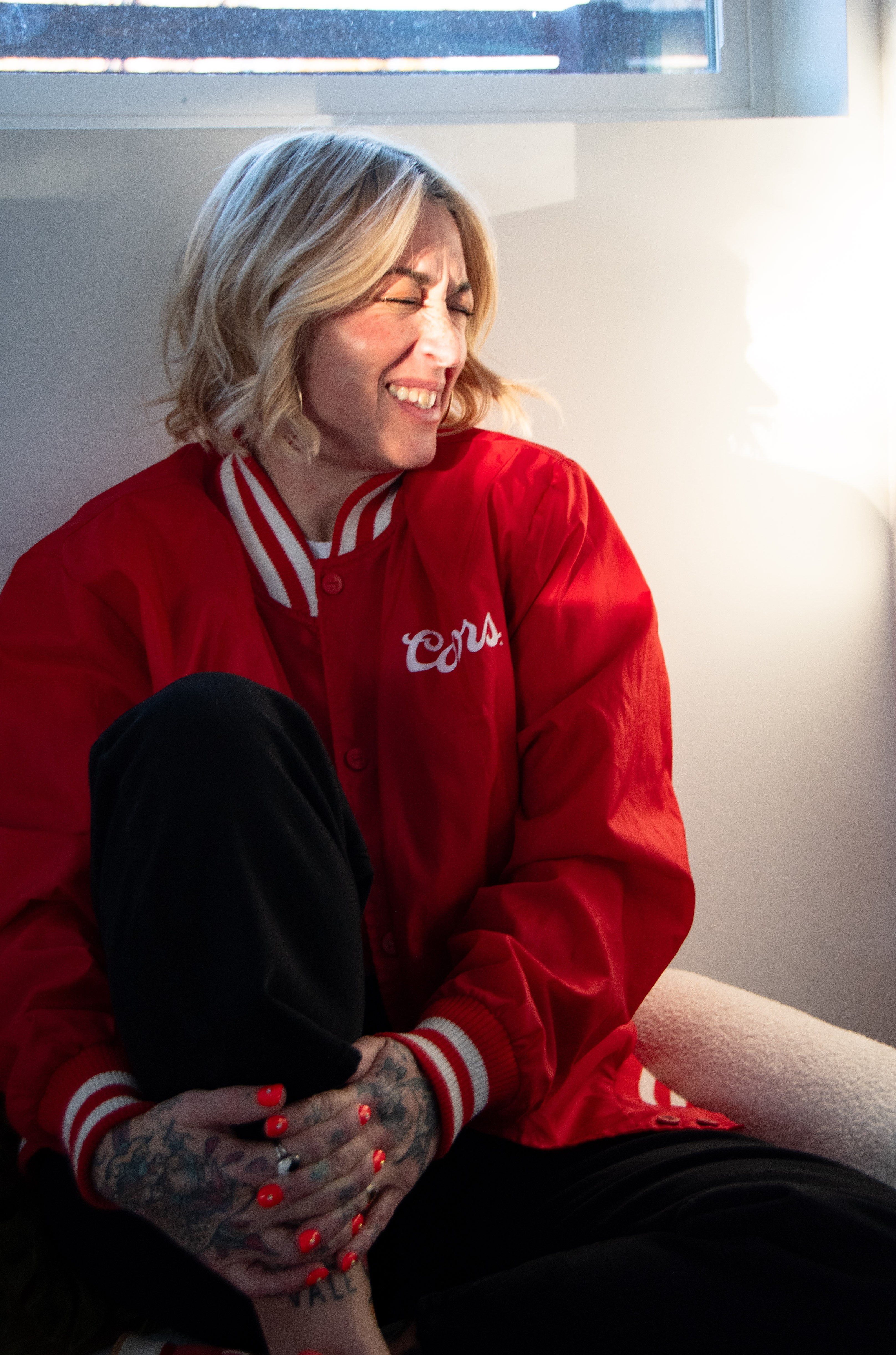 Woman with blonde hair smiling, wearing a red jacket with white stripes, sitting indoors near a window.