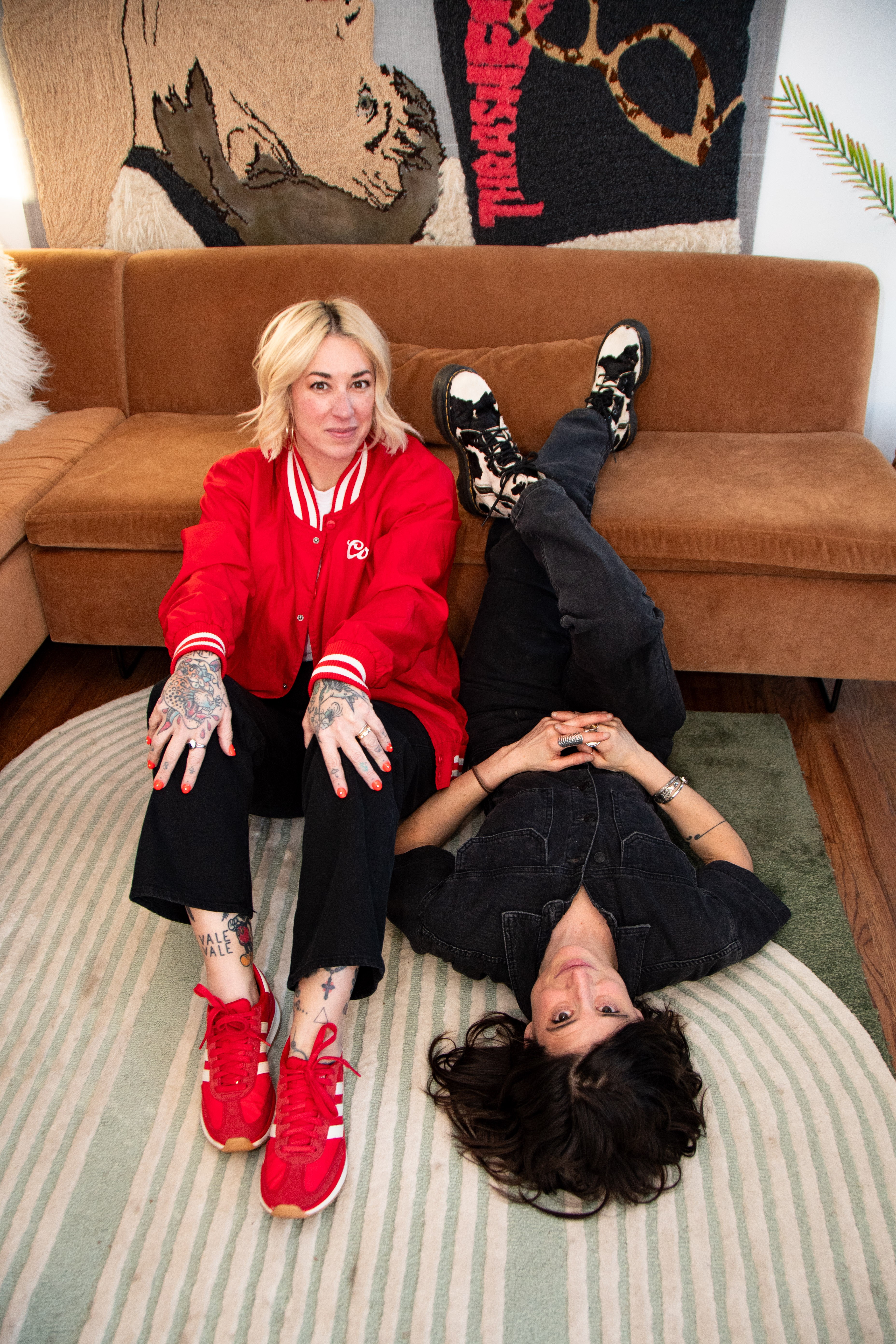 Two women in casual clothing on a striped rug, one sitting on a sofa, the other lying down with legs crossed.